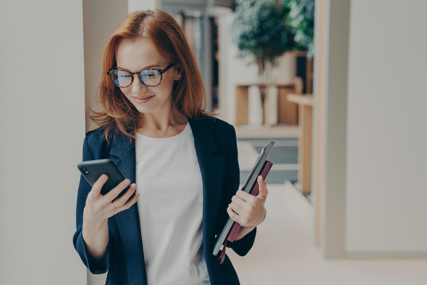 Elegant smiling woman office worker in eyeglasses using modern mobile phone in coworking space photo