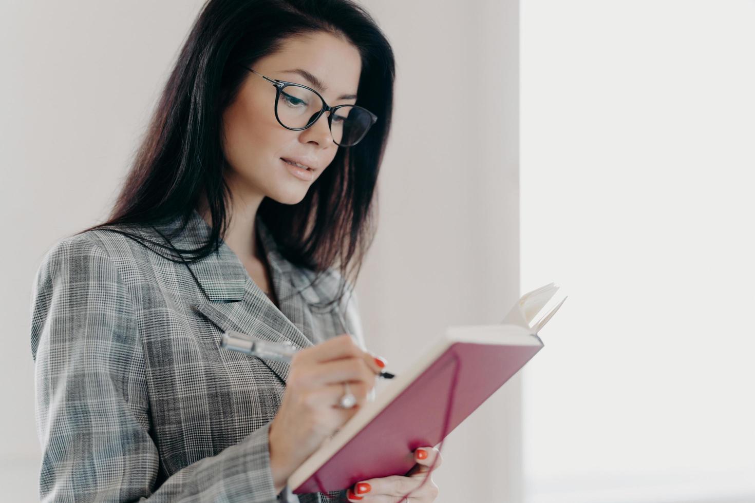 toma lateral de una mujer ejecutiva trabajadora con espectáculos, traje formal a cuadros, escribe información en el bloc de notas, hace una lista para hacer en el diario, apunta objetivos, posa en interiores, fondo blanco foto