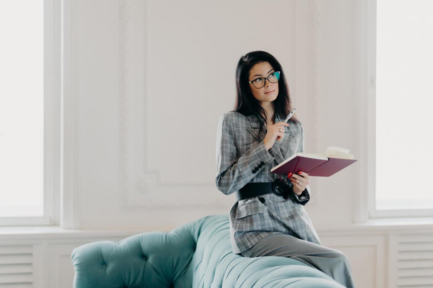 Serious businesswoman dressed in formal wear, concentrated into distance, ponders on creative ideas, writes down notes in notepad, wears spectacles, poses on back of couch in spacious light room photo