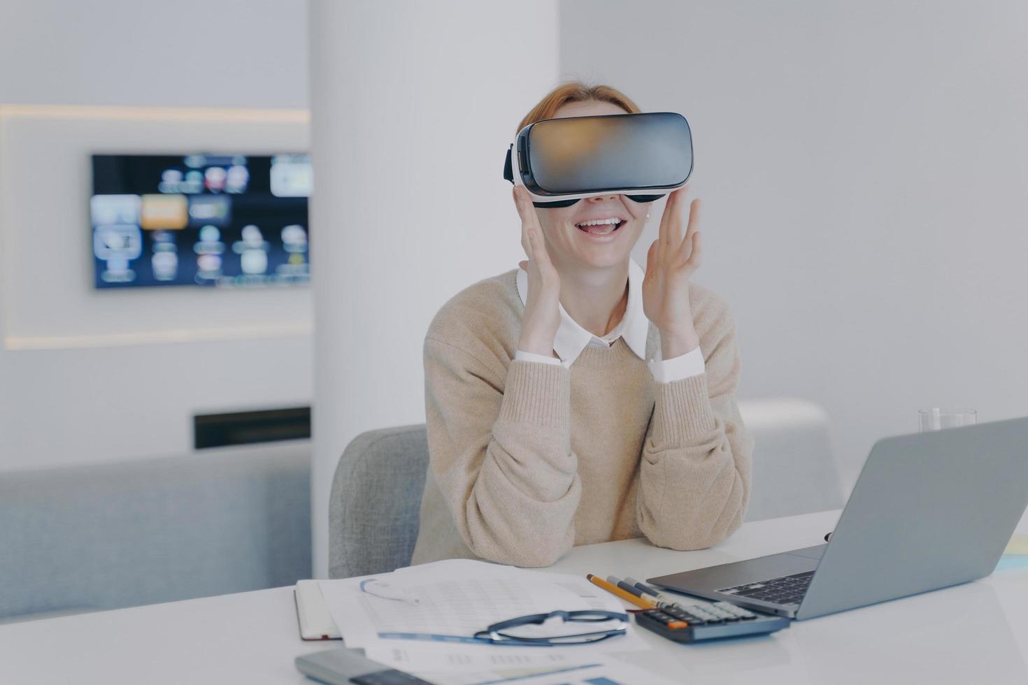 Excited girl in VR headset sitting at the desk in front of laptop. Working on project in cyberspace. photo