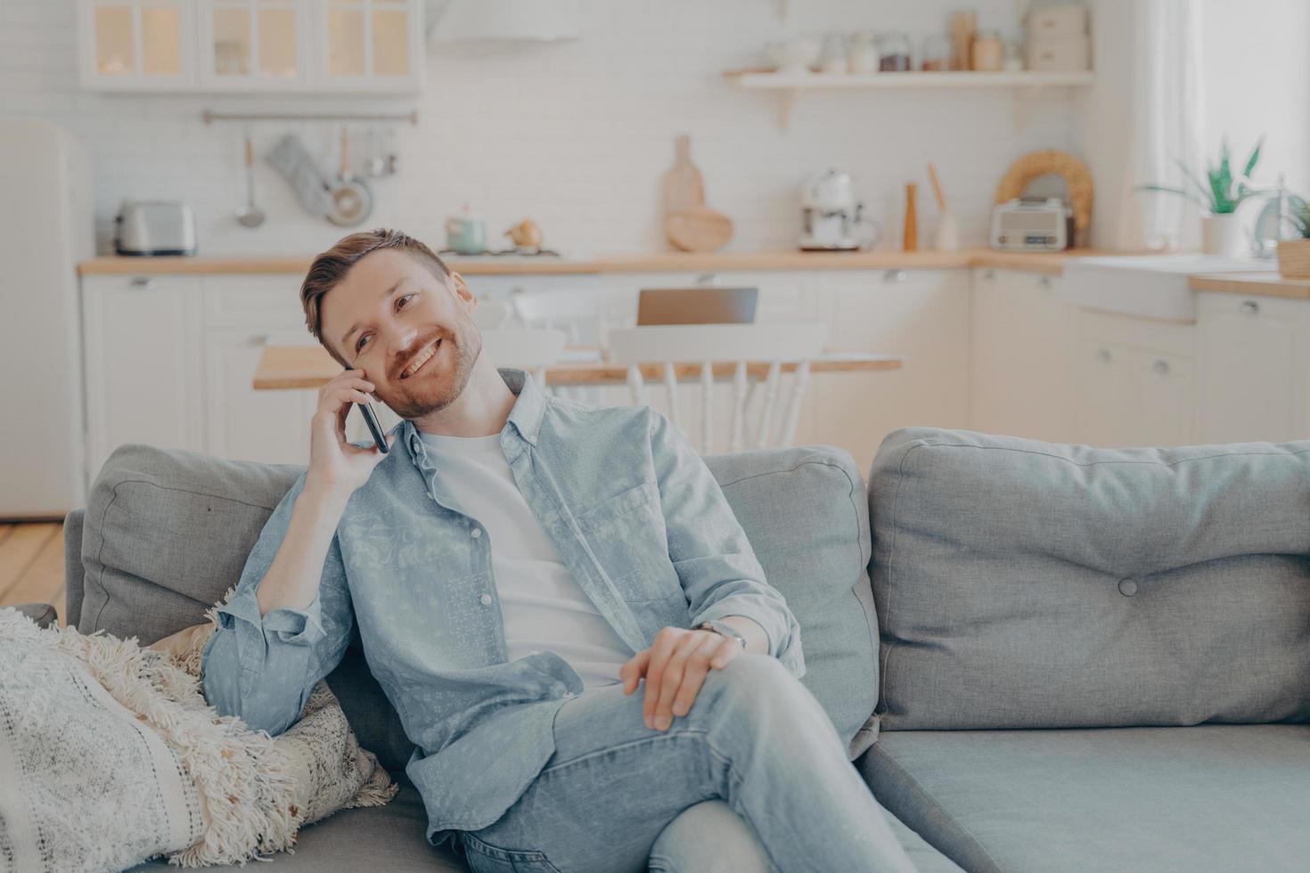 Smiling guy sits on couch at home having fun while talking on modern cellphone gadget photo