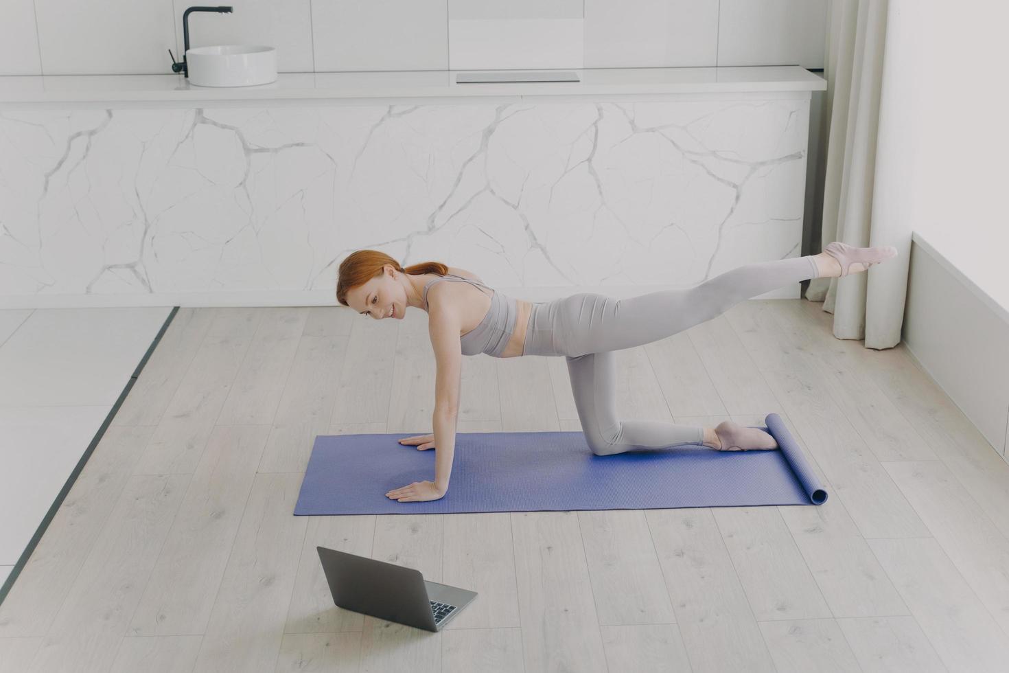 joven mujer europea feliz está haciendo ejercicio de tabla y mirando a la computadora portátil. entrenar en línea en casa. foto