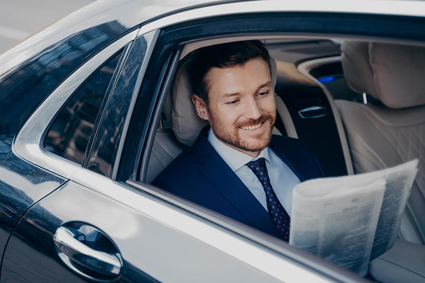 apuesto joven banquero con elegante traje formal de esmoquin lee el periódico en un auto de lujo foto