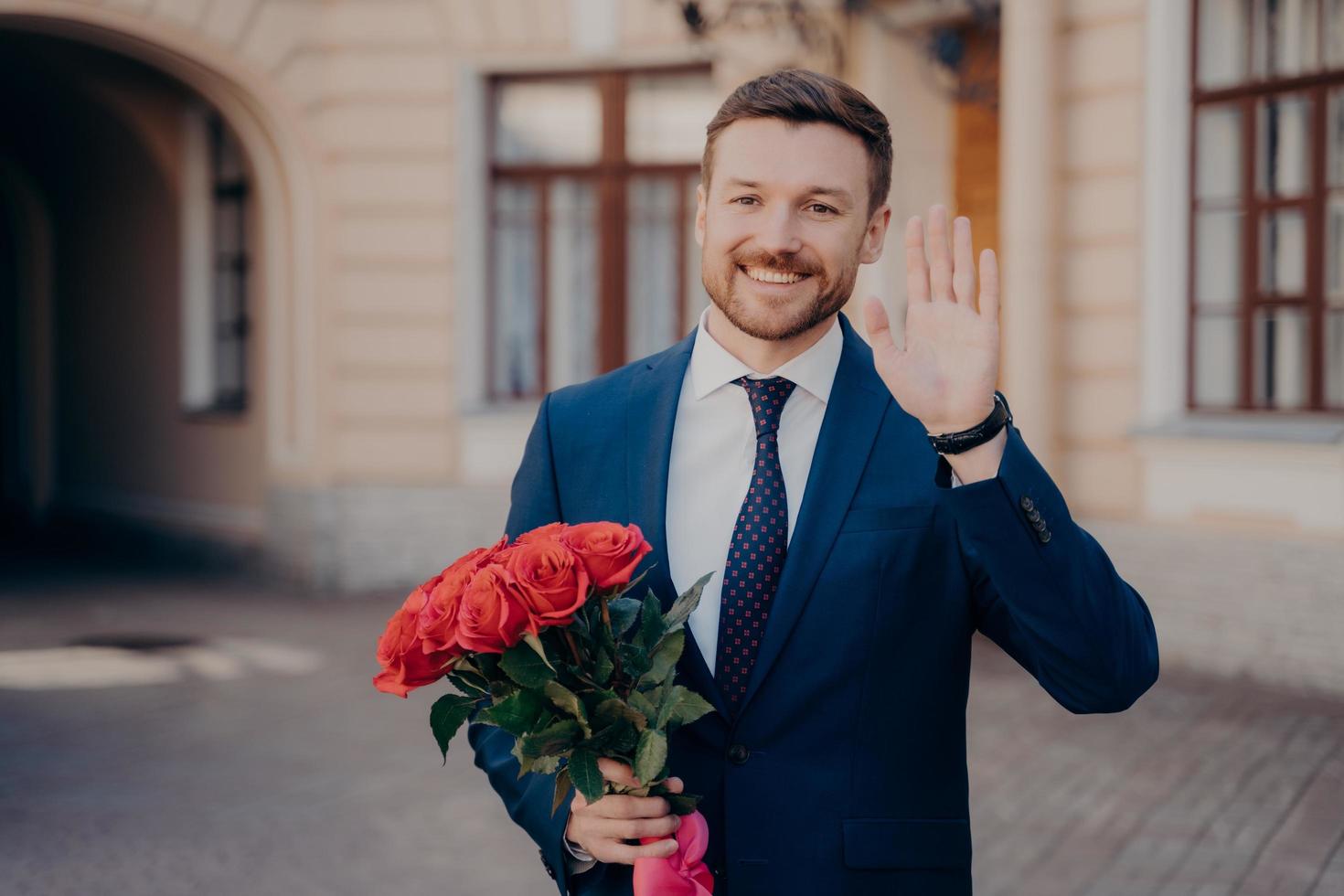 Attractive cheerful man in suit with bouquet of roses waving with hand while standing outdoors photo