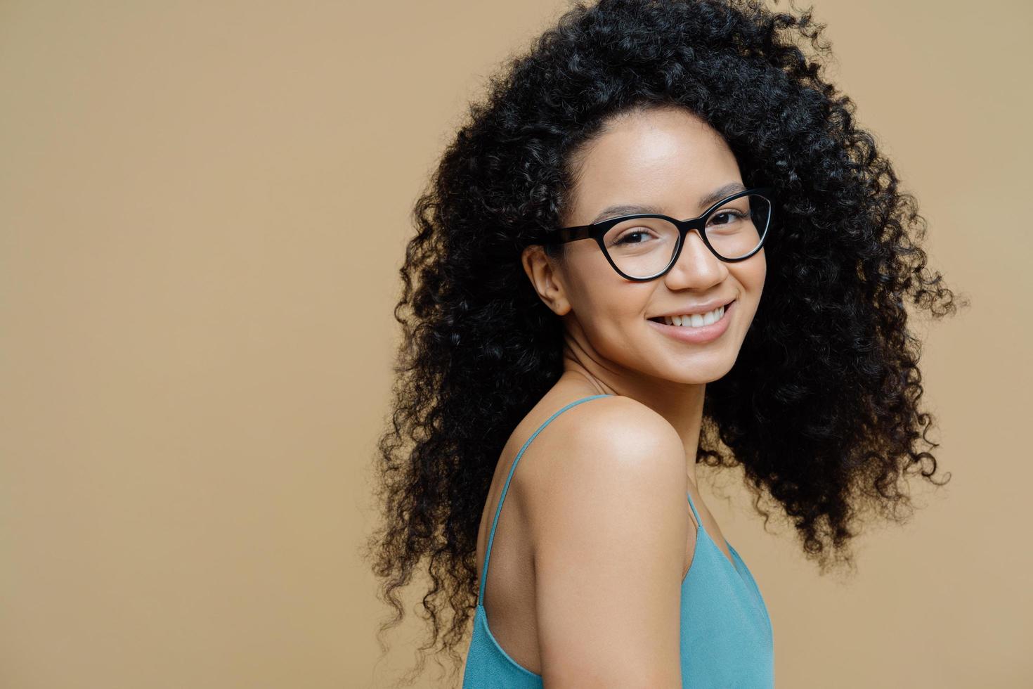 Magnetic pretty dark skinned woman stands sideways indoor, wears dress, shows bare shoulders, smiles gently, has curly bushy hair, looks through optical glasses at camera, isolated on beige wall photo