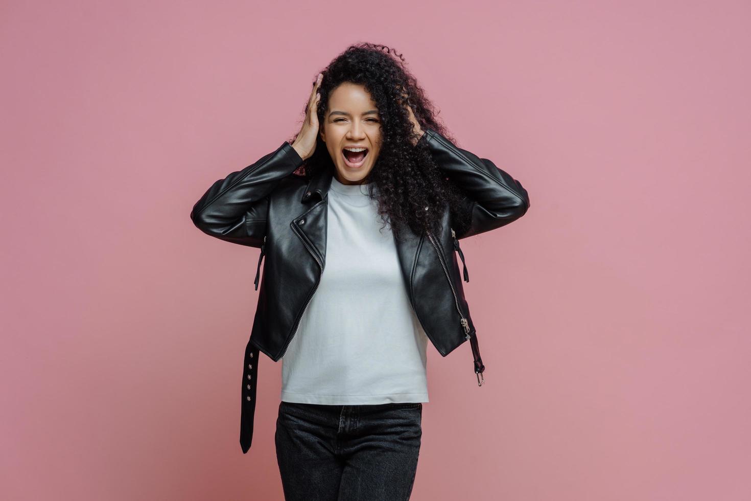 Emotional crazy adult woman grabs head and shouts loudly, hears very loud music, keeps mouth opened, dressed in stylish leather jacket, isolated on pink background. People and emotions concept photo