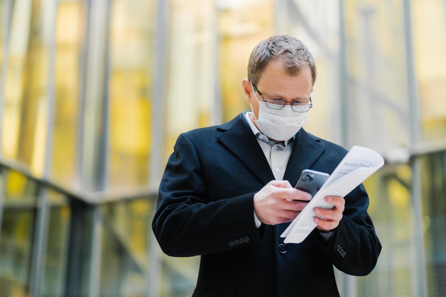 concepto de pandemia de coronavirus. foto de un gerente masculino enfocado en un dispositivo de teléfono inteligente, envía mensajes de texto, usa anteojos y máscara médica, sostiene papeles, camina por la calle durante la cuarentena, la pandemia