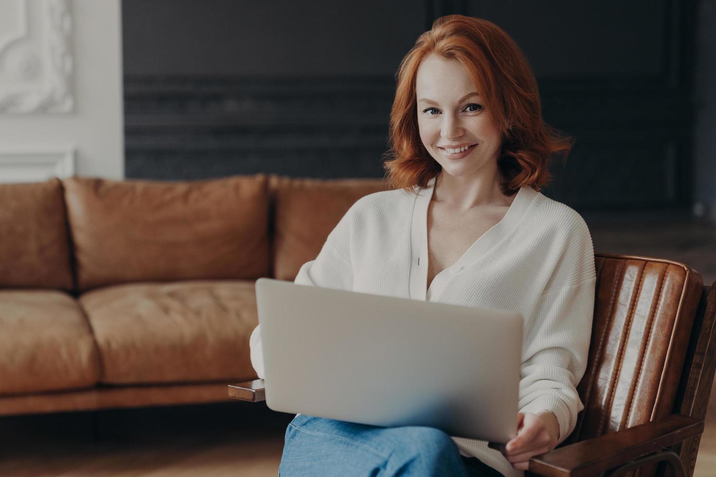 hermosa joven pelirroja con puente blanco sentada en un sillón con computadora portátil, se prepara para una entrevista en línea con un nuevo empleado, un gran sofá cómodo en el fondo. trabajar lejos de casa foto