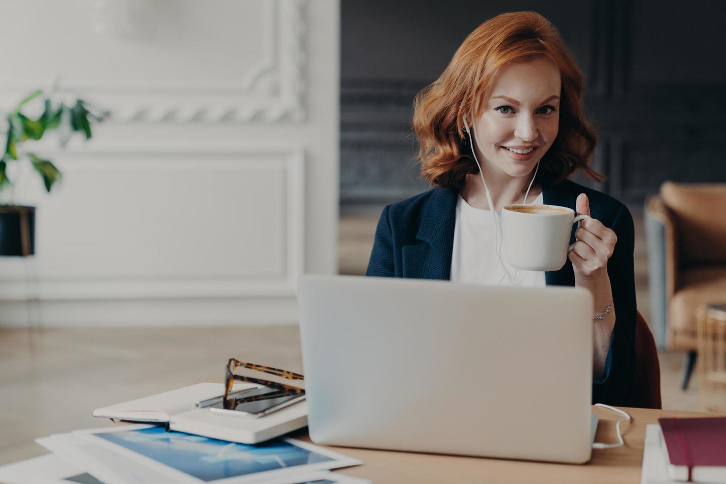 Happy young redhead woman watches training webinar on laptop computer, browses information for project, drinks coffee or espresso, poses at desktop, uses earphones for e learning, autodidact photo
