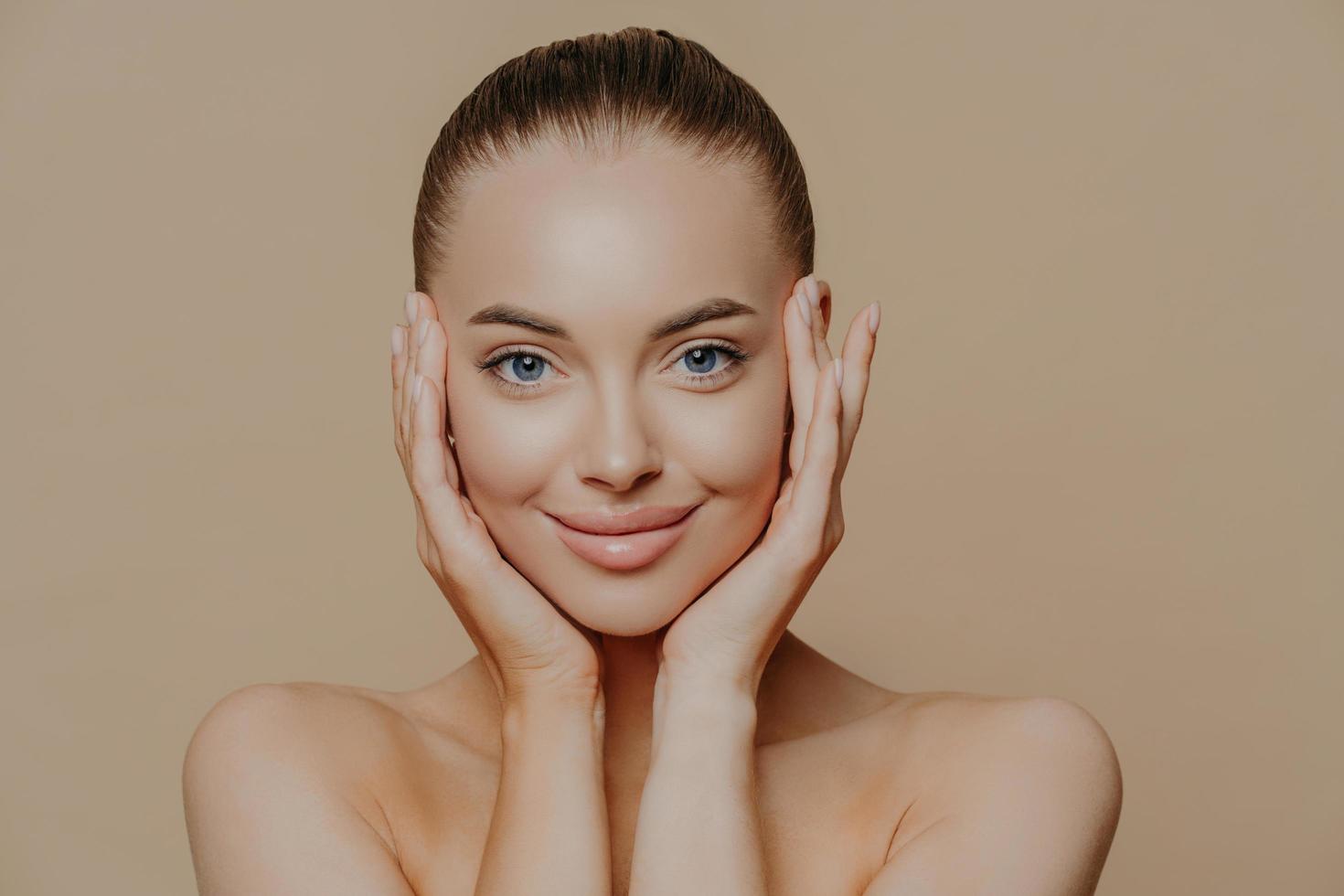 Close up portrait of blue eyed glad woman with dark combed hair, touches face gently, has healthy perfect skin, bare shoulders, enjoys beauty procedures, models indoor. Skin care and wellness photo