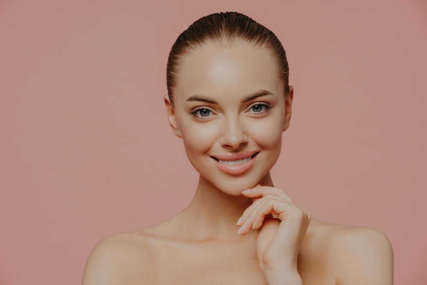 Studio shot of pretty charming woman touches chin, has healthy glowing skin, cares about complexion, stands naked indoor, has cosmetology procedure, isolated on pink background. Beauty concept photo