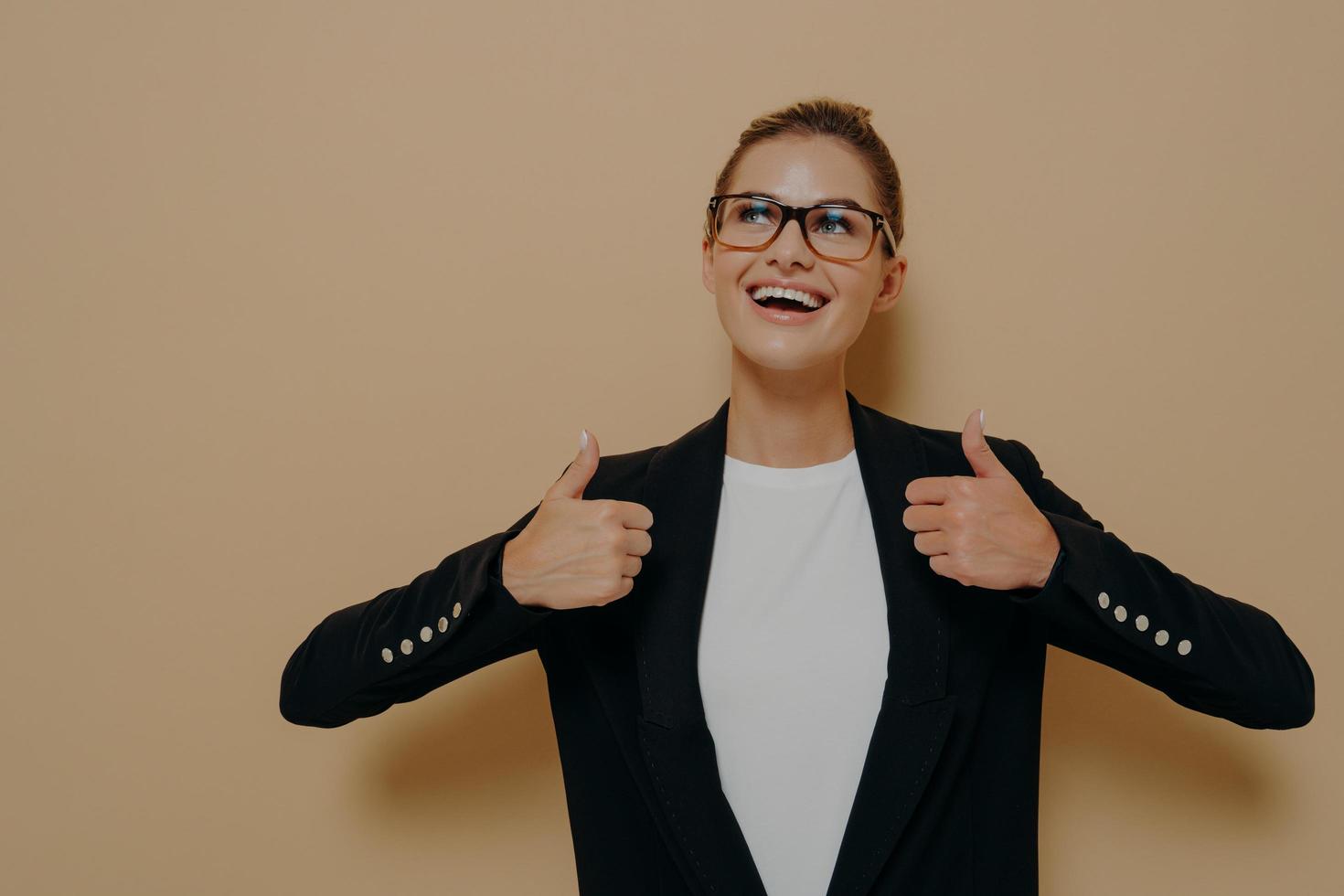 Positive female customer in eyeglasses with broad smile showing thumbs up with both hands photo