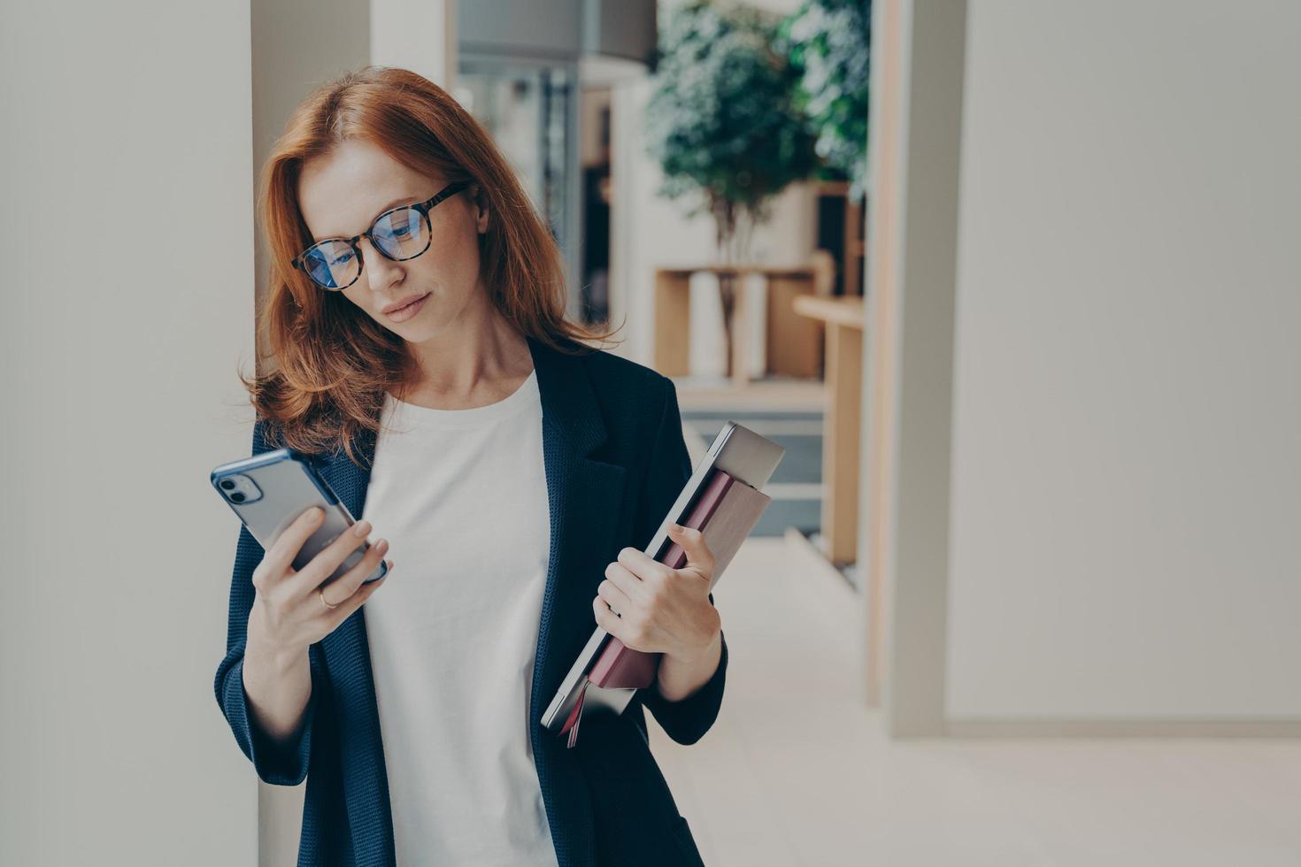 Redhead business woman with serious look in eyewear using cell phone while standing in office photo