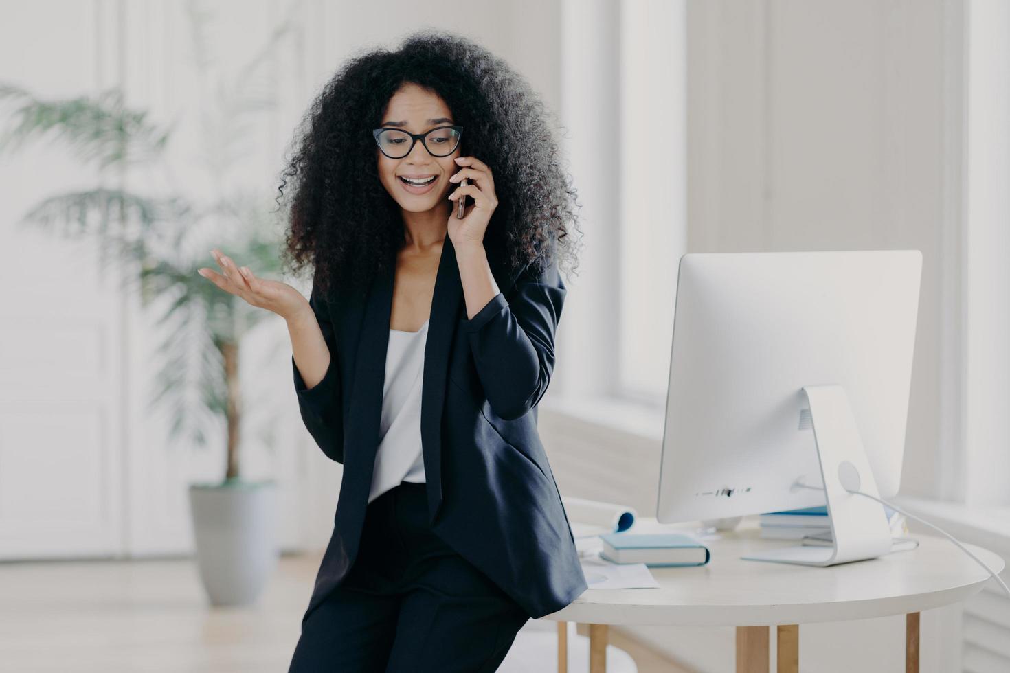 Photo of puzzled prosperous businesswoman calls partner, raises palm, holds mobile phone, wears spectacles and formal black outfit, tries to solve problem at work, discusses something actively