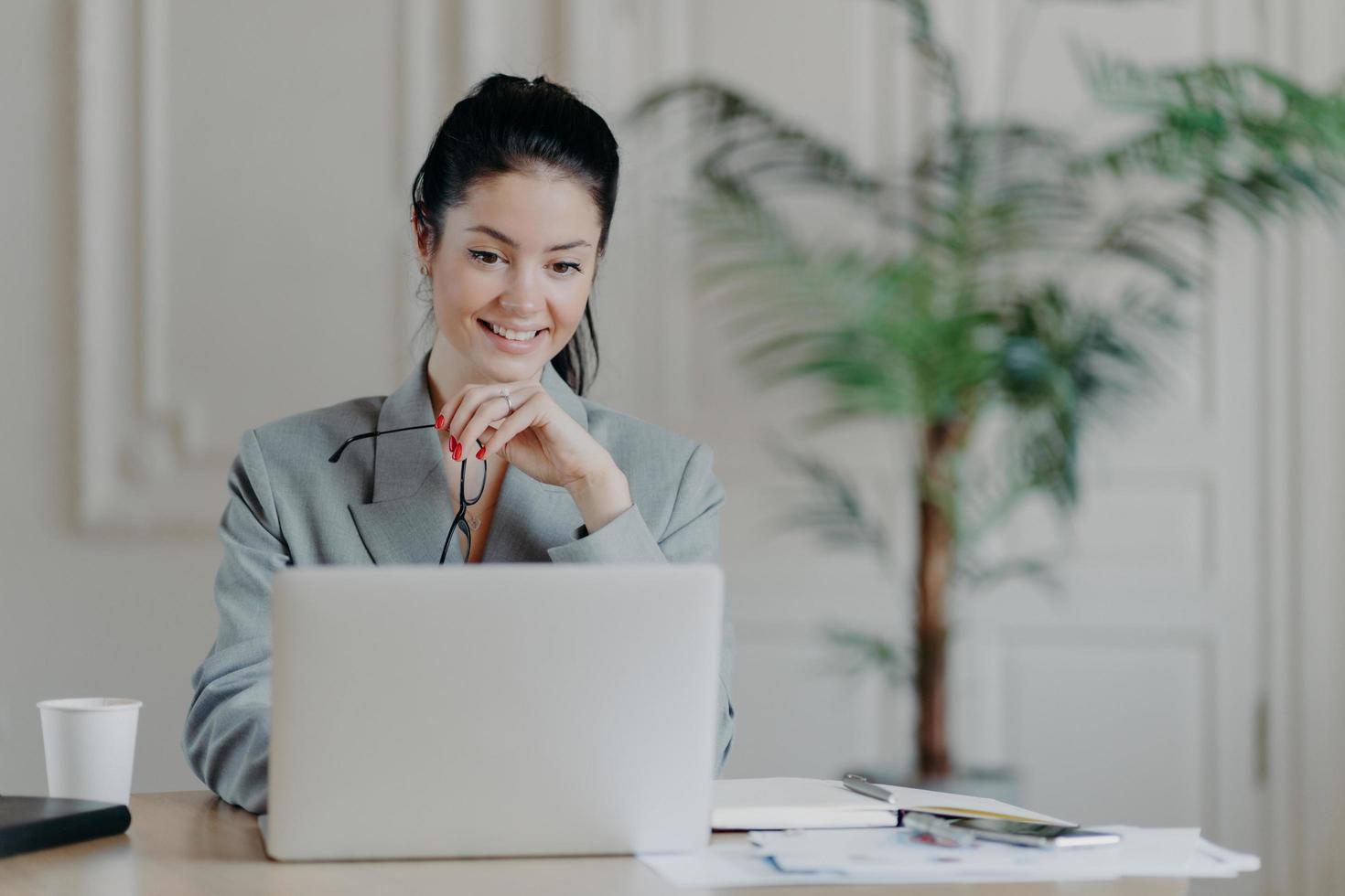 Cheerful young professional woman searches update notification for laptop computer, takes off glasses, dressed in formal clothes office interior. Freelancer works remotely searches ideas for blog site photo