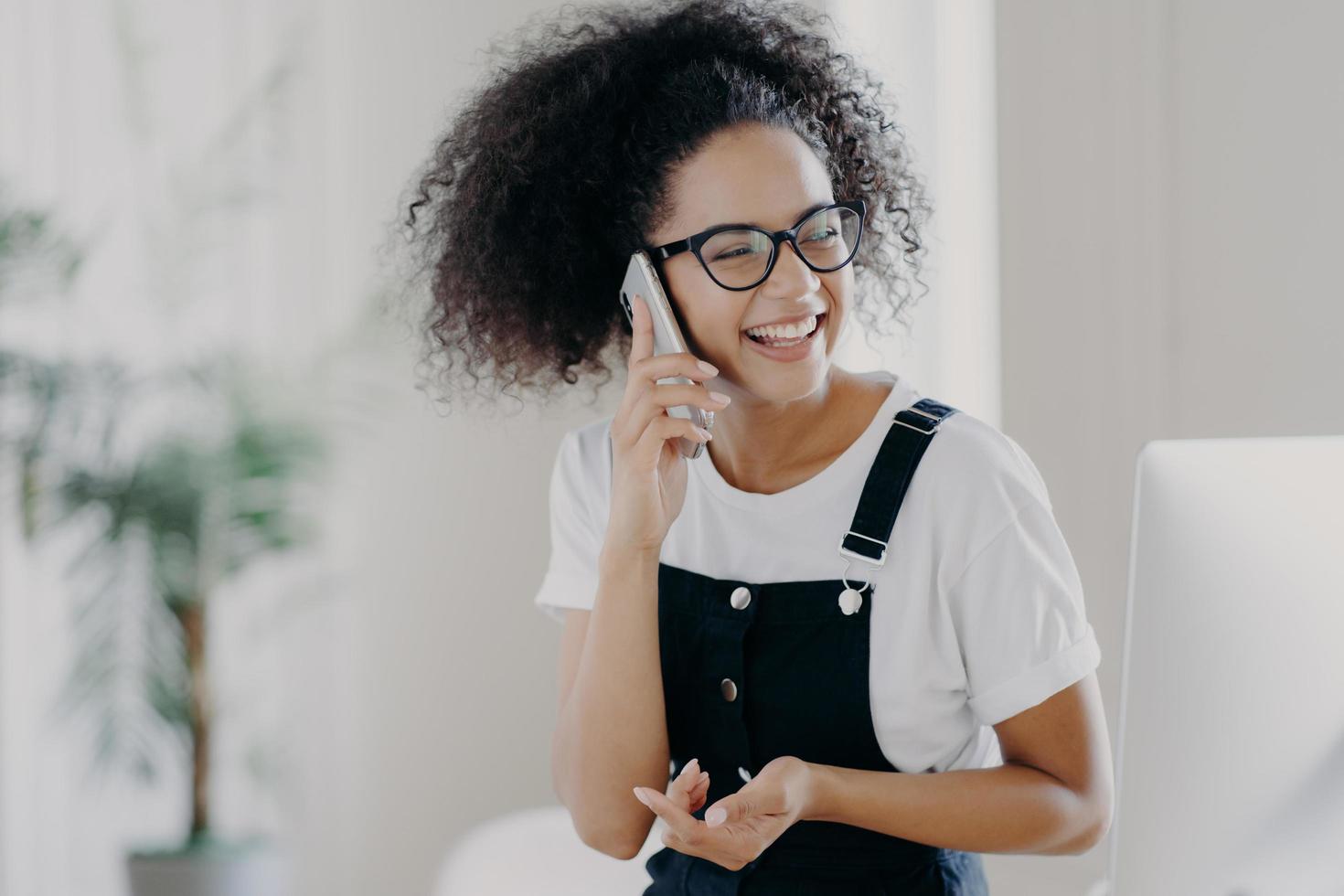 Photo of overjoyed female discusses something pleasant via cell phone, holds cellular near ear, being in good mood, wears glasses and casual clothing, poses in spacious room, blurred background