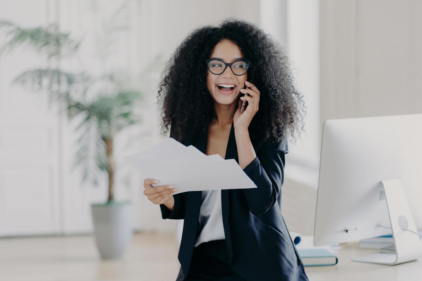 Pleased curly haired female director dressed in formal outfit, holds documents, calls via cell phone, stands near workplace with computer, shares opinion about startup, makes business report photo