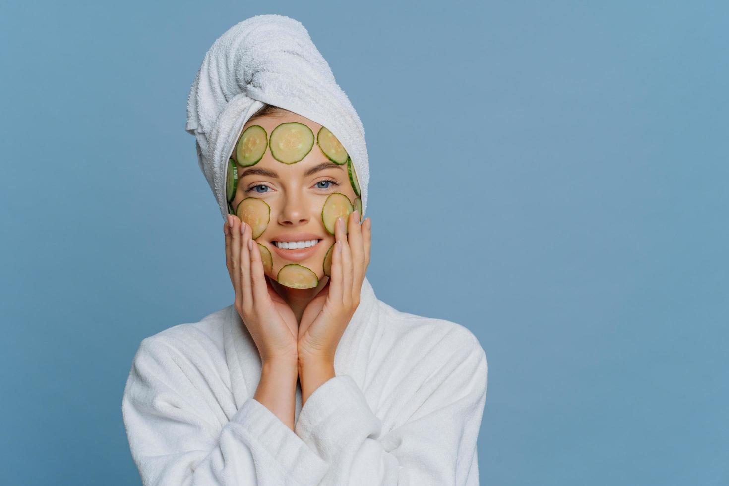 una joven complacida aplica rodajas de pepino en la cara se somete a procedimientos de belleza y hace una máscara facial para rejuvenecer la piel vestida con una bata de baño blanca y una toalla envuelta en la cabeza aislada en la pared azul foto