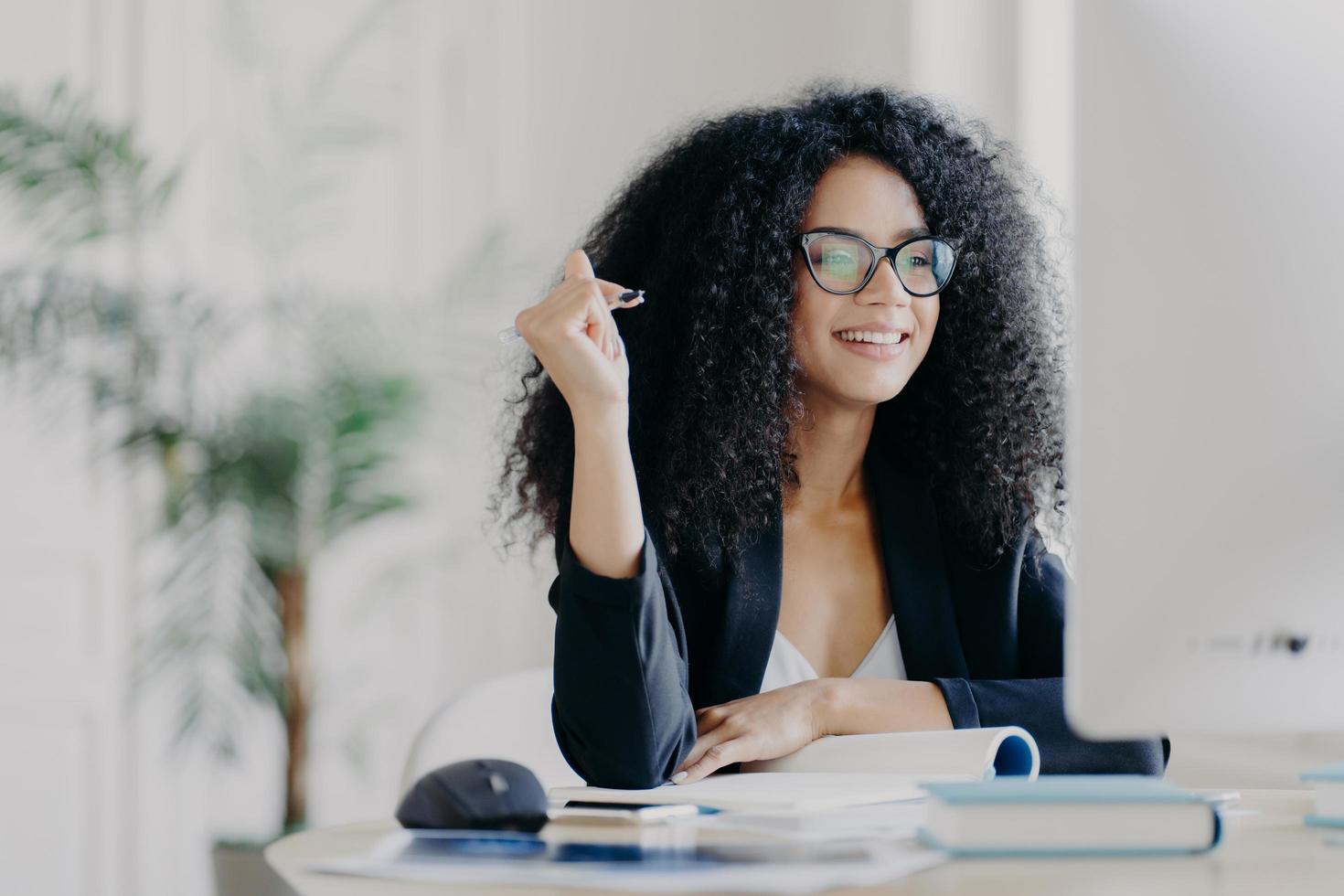 una linda mujer afroamericana mira positivamente la pantalla de la computadora, escribe información de Internet, usa anteojos ópticos y traje negro, se sienta en el escritorio con las cosas necesarias para el trabajo foto