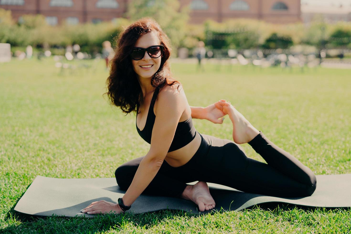 Outdoor shot of sporty smiling woman stretches legs on karemat smiles positively wears activewear has fitness workout for flexibility does stretching exercise enjoys fresh air and green lawn. photo
