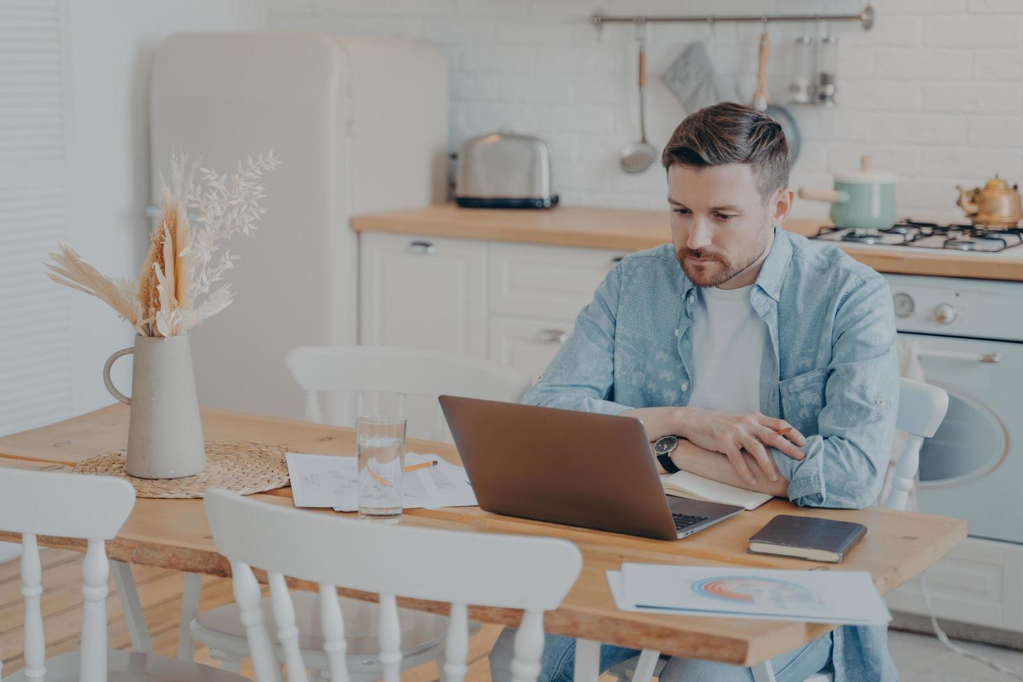Focused young freelancer working on laptop while sitting by kitchen table photo