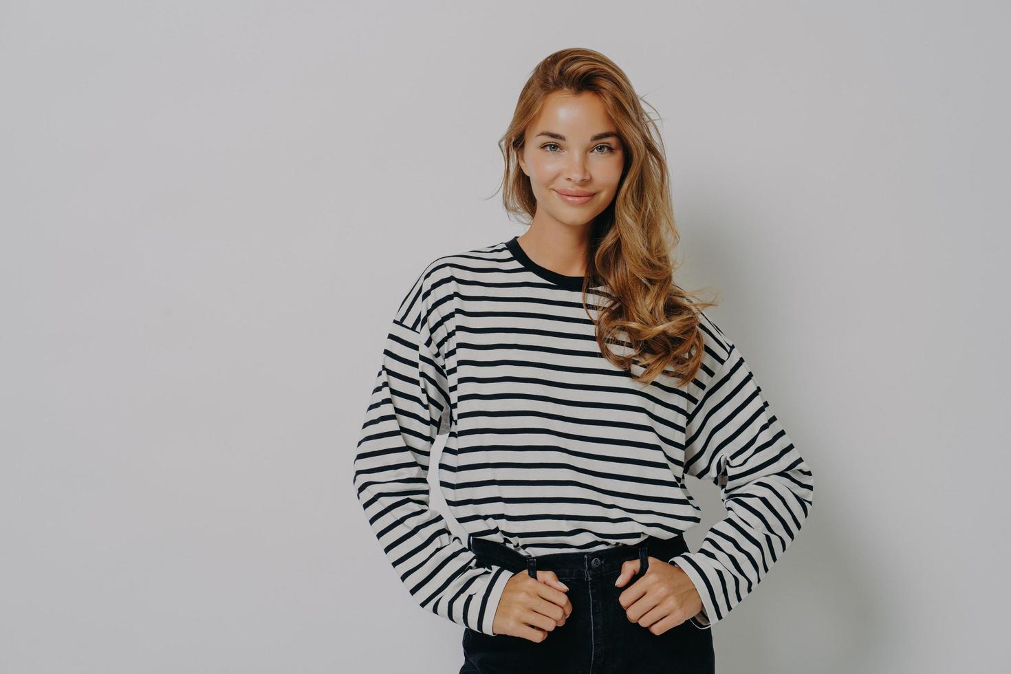 Young confident charming woman in striped blouse and casual jeans posing with hands on waist photo