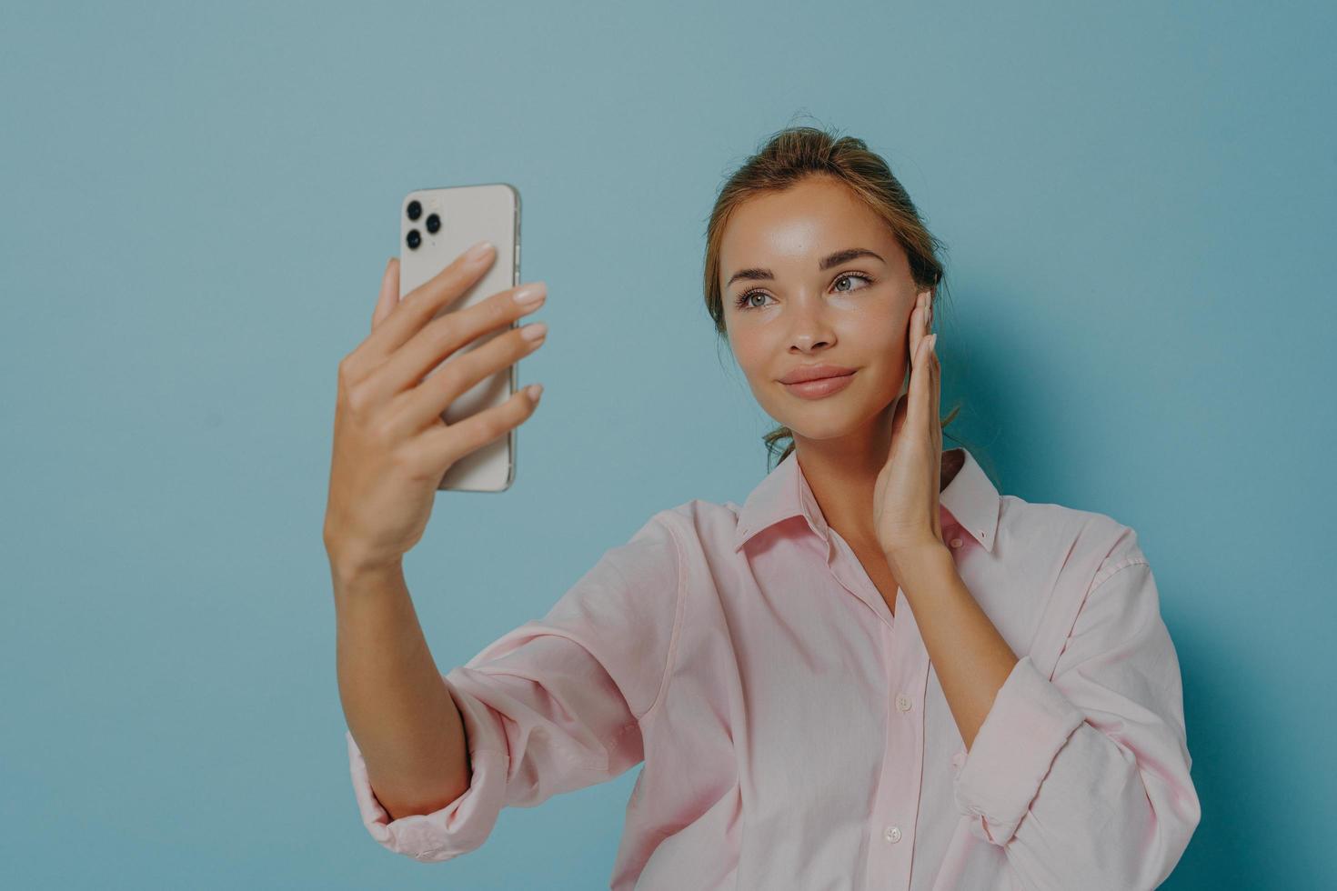 Beautiful woman with tender expression looks at smartphone camera takes selfie of herself wears photo