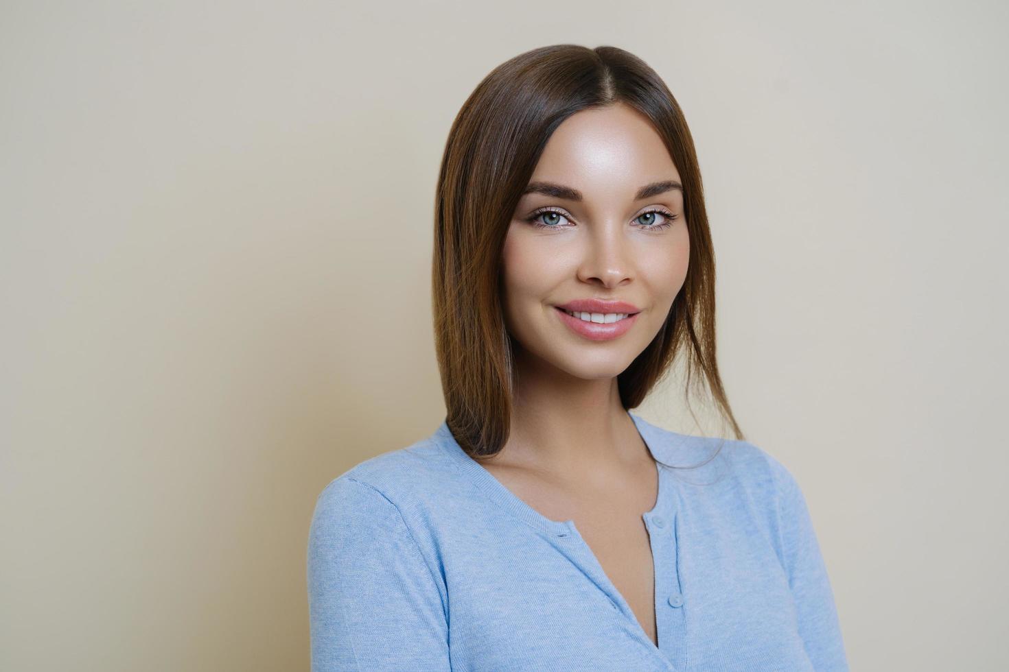 mujer joven morena con piel de belleza saludable, sonríe suavemente, muestra dientes blancos y perfectos, usa maquillaje mínimo, se viste con un jersey casual, mira la cámara y sonríe, aislada en un fondo beige. foto