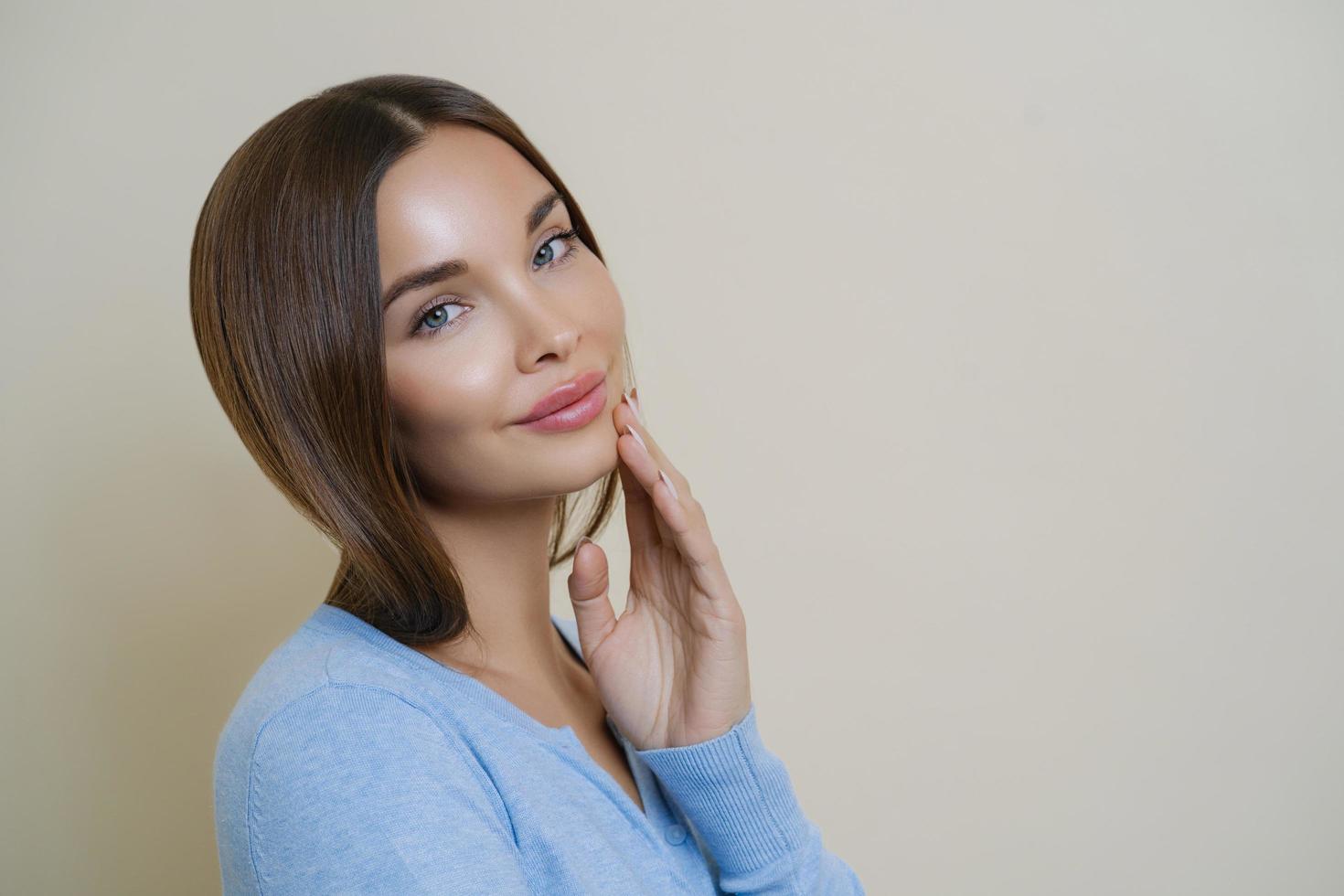 concepto de personas, mujeres, belleza y cuidado personal. Una encantadora mujer europea de cabello oscuro toca la mejilla suavemente, mira directamente a la cámara, posa para hacer una foto en la portada de la revista, tiene una apariencia atractiva