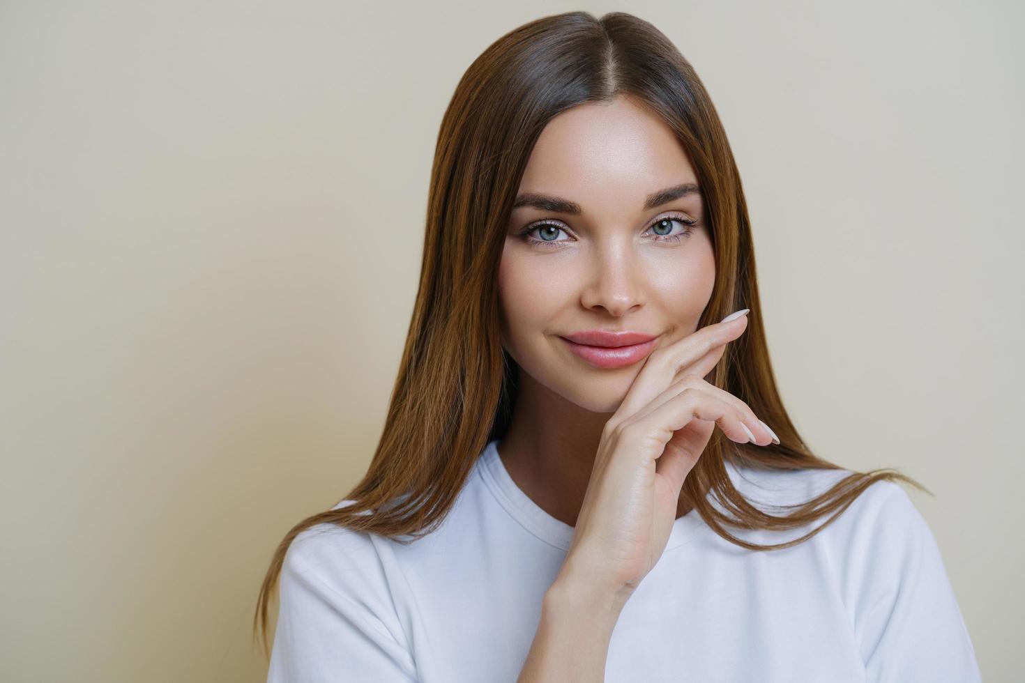 Close up shot of charming dark haired woman keeps hand on cheek, dressed in casual white t shirt, has minimal makeup, poses indoor isolated over beige background. People, beauty, wellness concept photo