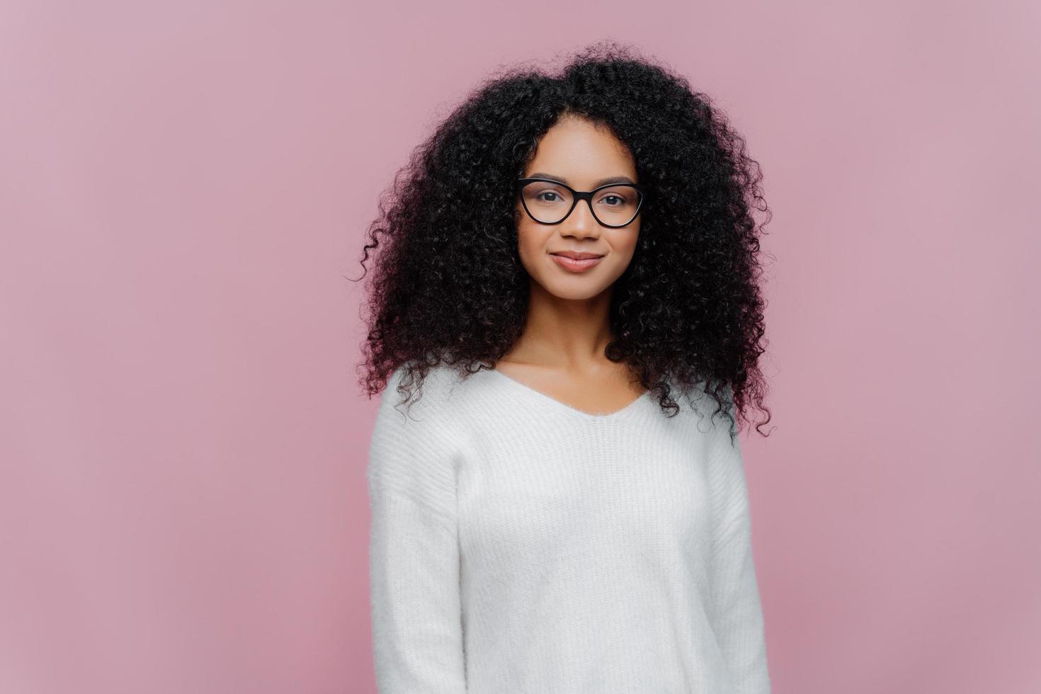 foto de media longitud de una atractiva mujer afroamericana mira a través de gafas transparentes, suéter blanco, tiene una expresión seria y tranquila, posa contra la pared violeta del estudio. concepto de expresiones faciales