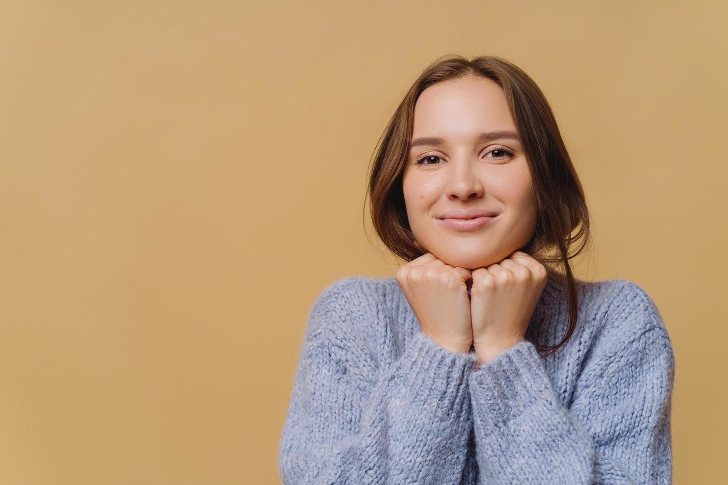 Lovely dark haired woman with healthy skin, keeps both hands under chin, wears oversized sweater, looks directly at camera, isolated over brown studio wall. People, human facial expressions. photo