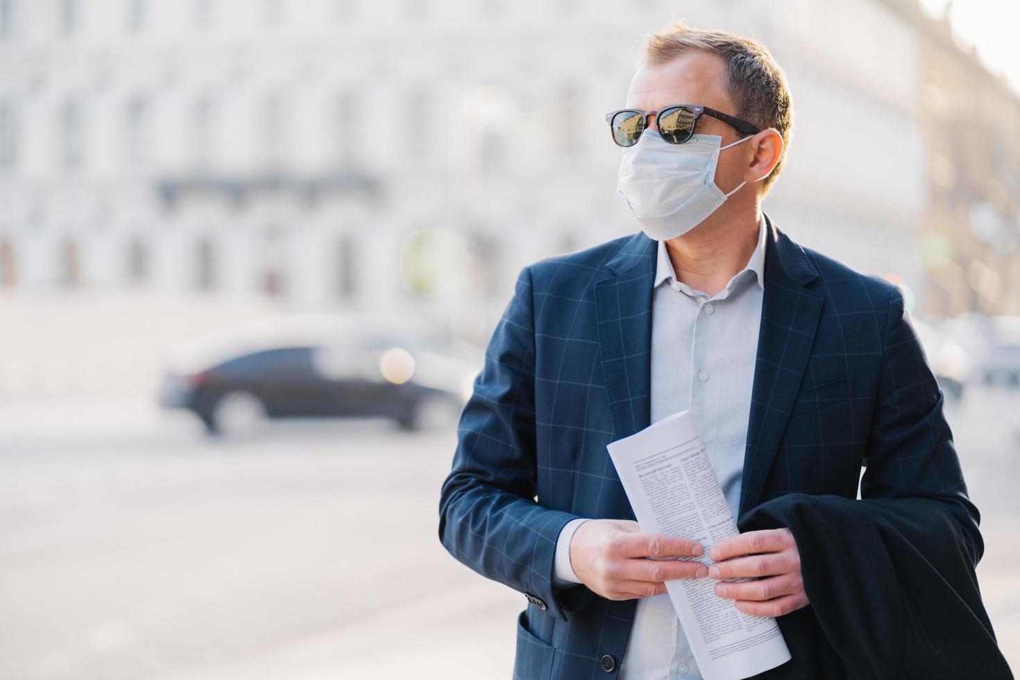 Covid-19. Man worker dressed in formal wear, holds newspaper in hands, poses at street, wears surgical mask, avoids crowded street not to catch virus, poses against blurred background with transport photo