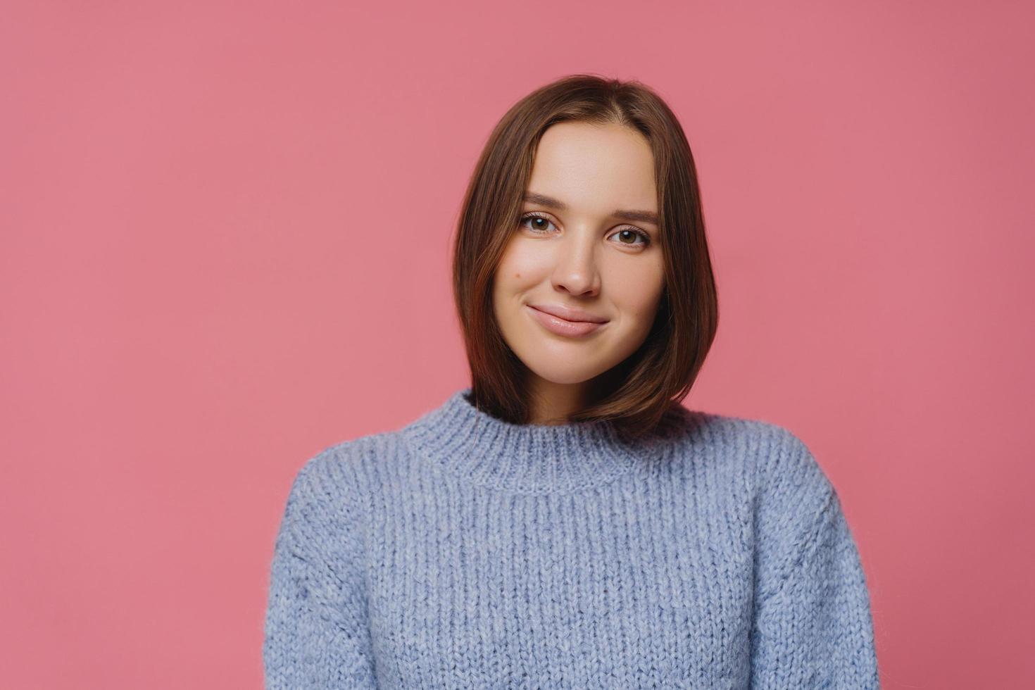 Horizontal shot of pleasant looking dark haired woman in knitted sweater looks tenderly at camera, enjoys winter day and coziness, poses against pink background. Feminity and emotions concept photo