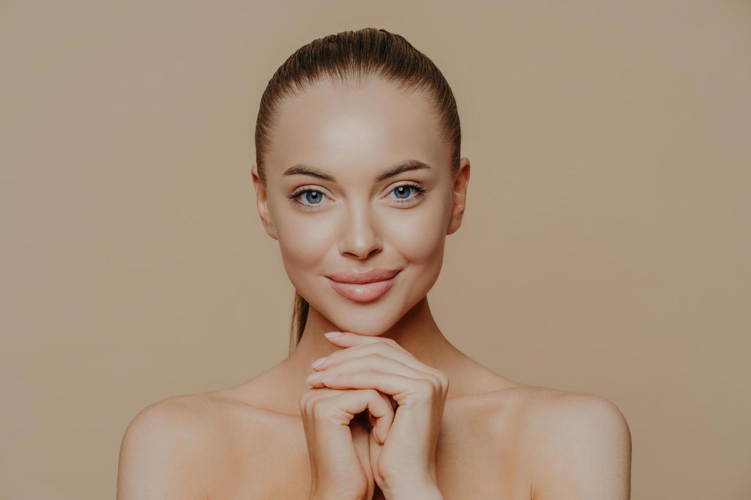 Young pretty woman with beauty skin and combed hair, keeps hands under chin, has healthy complexion after face purification, soft well cared body, isolated over studio wall. Facial treatment photo