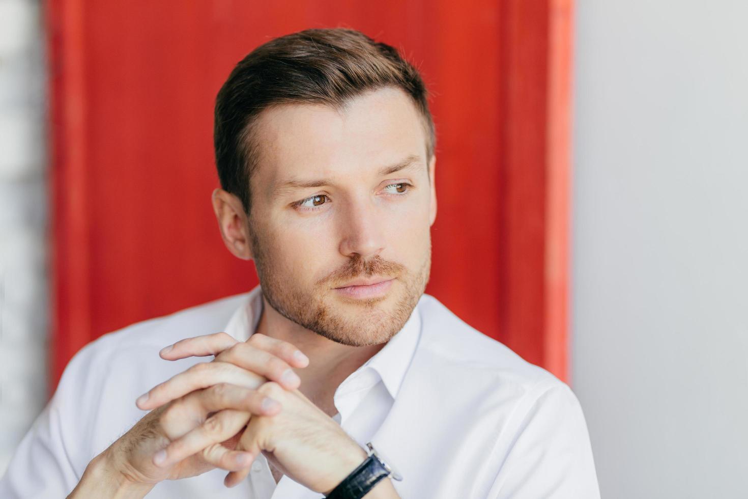 Horizontal shot of pensive handsome young male corporate worker holds hands together, looks aside, being deep in thoughts after meeting, poses over red background. Man leader owns big company photo