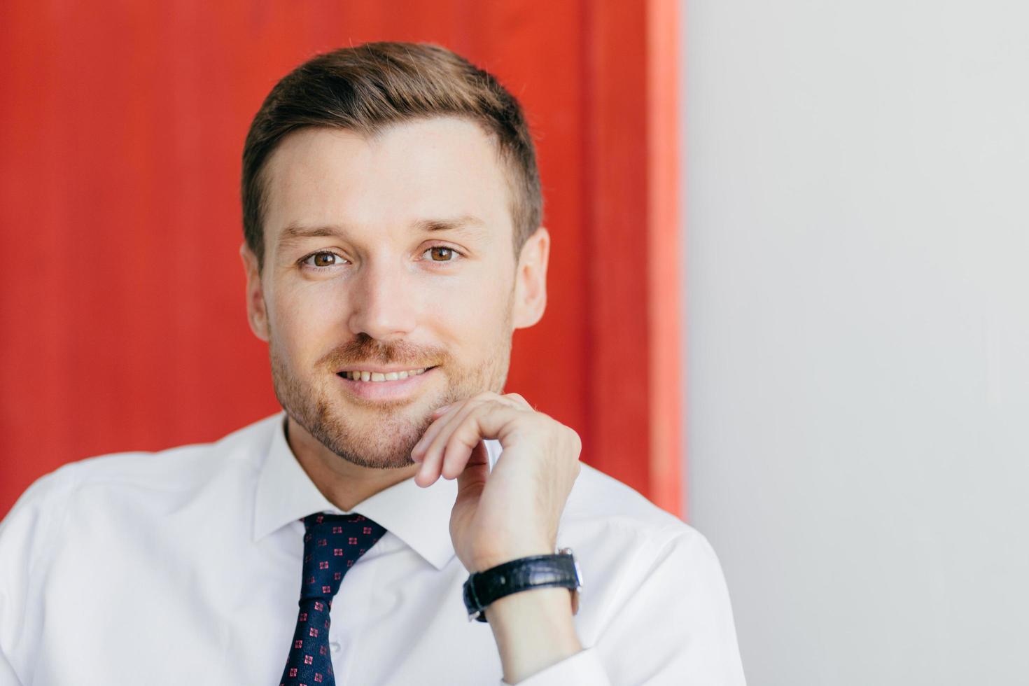 Prosperous young male entrepreneur dressed in white shirt, looks positively at camera, being confident in his success, isolated over red and white background, thinks about starting new career photo