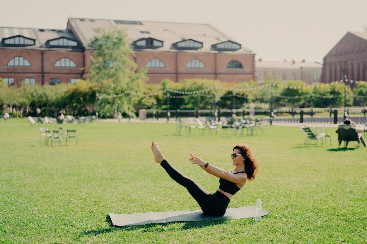 concepto de pilates fitness y pérdida de peso. mujer joven morena positiva tiene entrenamiento al aire libre levanta poses de piernas en el entrenamiento regular de karemathas usa ropa deportiva gafas de sol estira los músculos. foto