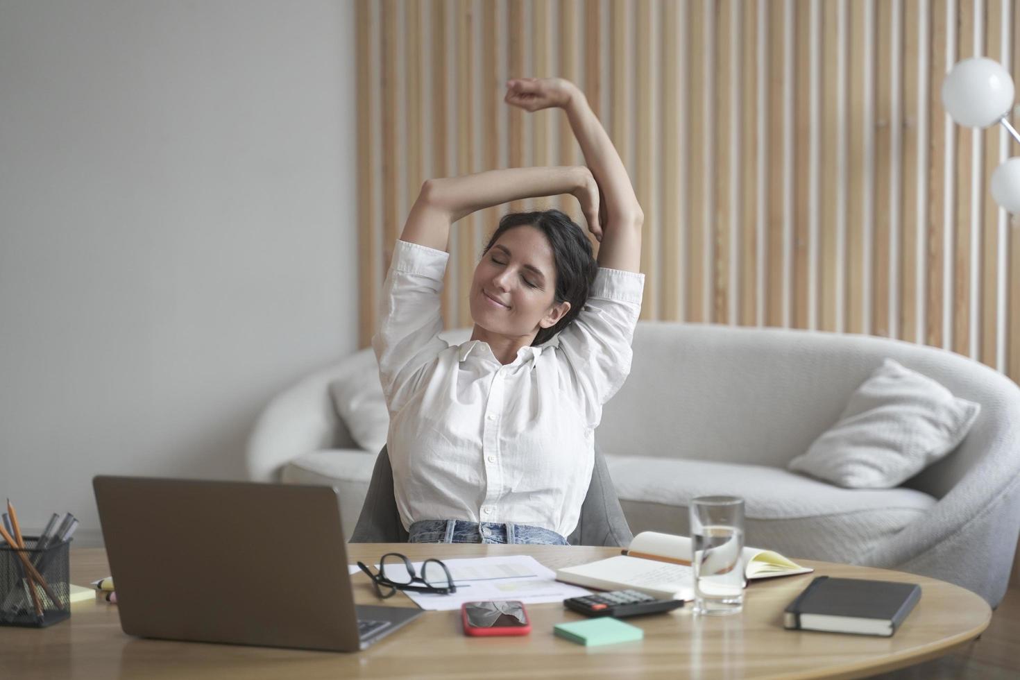 Tired italian lady remote employee stretching arms while sitting at desk with laptop photo