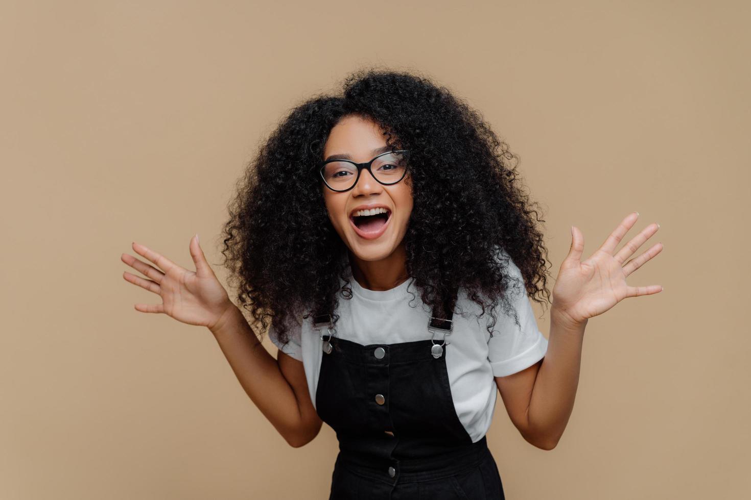 Overjoyed African American woman laughs out, raises palms, being in high spirit, wears transparent glasses, casual t shirt and overalls, poses against brown background, feels happiness and pleasure photo