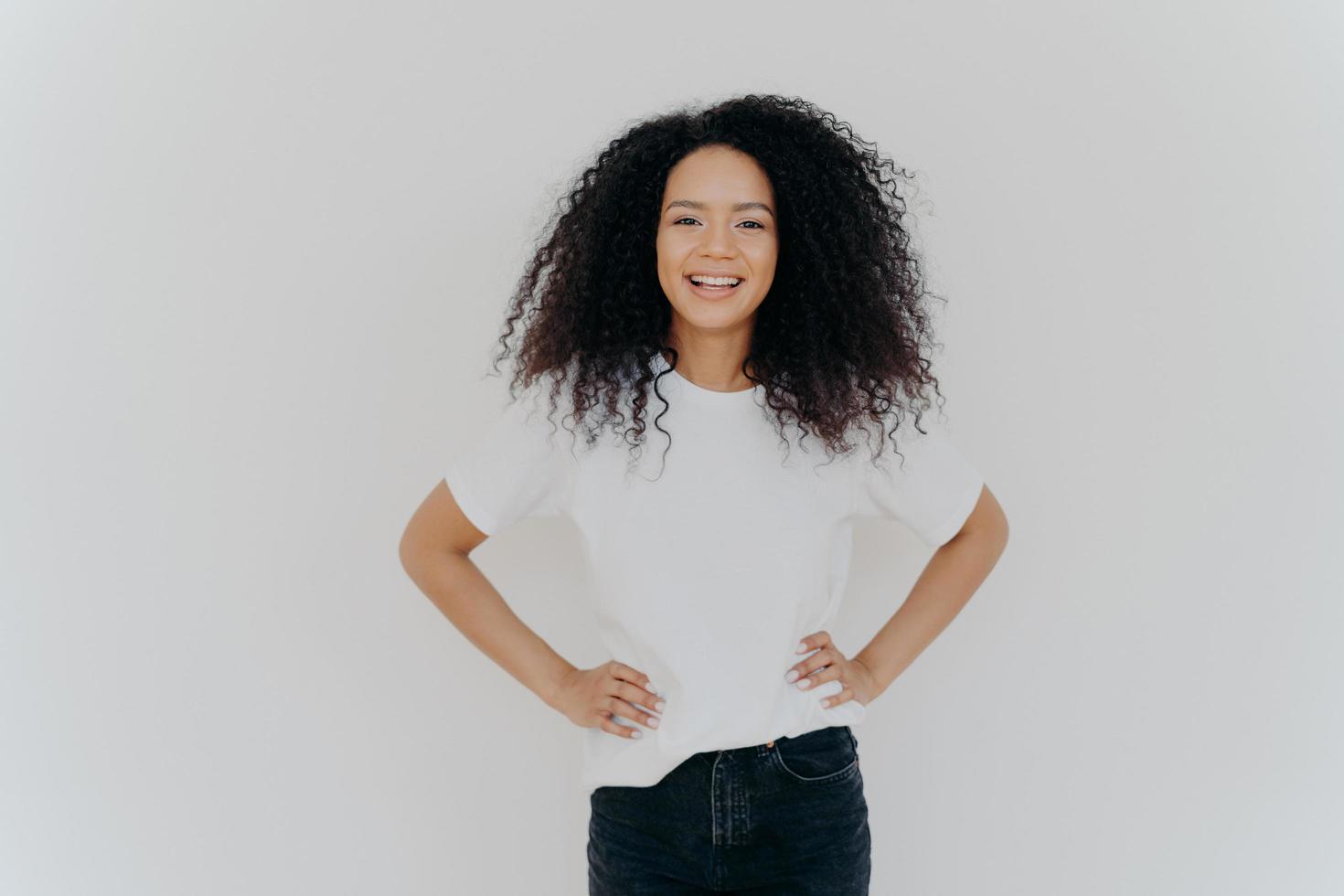 foto de media longitud de una hermosa y alegre niña milenaria parada con una expresión de cara feliz contra el fondo blanco del estudio, mantiene ambas manos en la cintura, tiene el pelo rizado, maquillaje mínimo, figura delgada