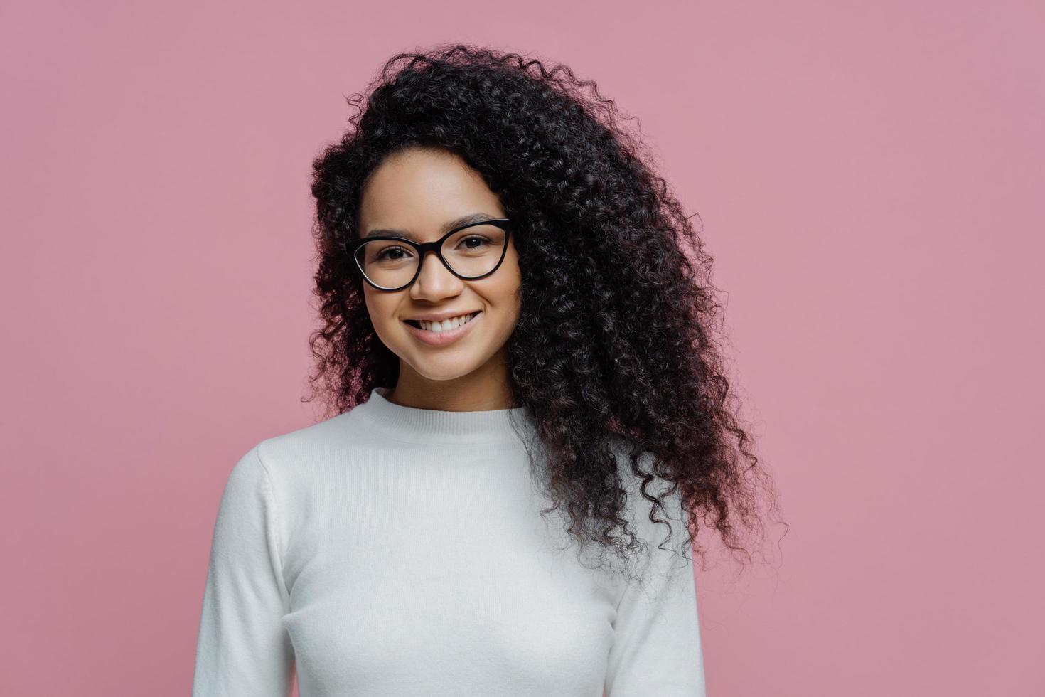 tiro en la cabeza de una encantadora mujer de cabello rizado con piel sana y sonrisa con dientes, vestida con un puente blanco, usa anteojos transparentes, aislada sobre un fondo rosado. personas, expresiones faciales y emociones foto