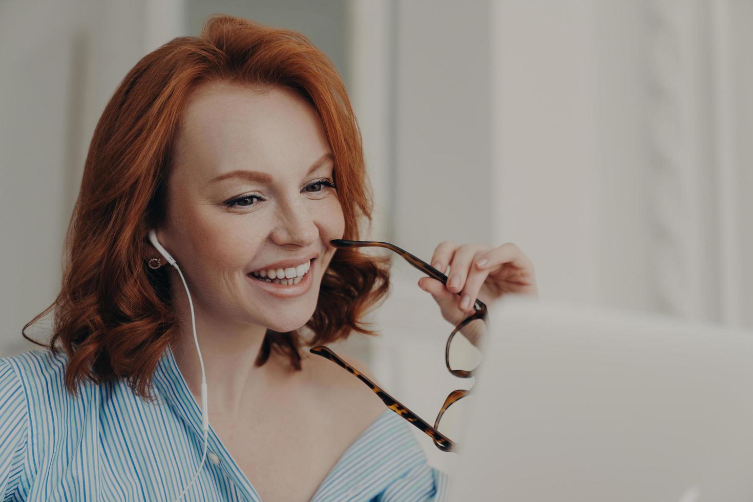 Beautiful cheerful redhead woman uses laptop computer for online conference, talks with colleagues distantly, holds spectacles, discusses working issues, works freelance from home. Technology concept photo