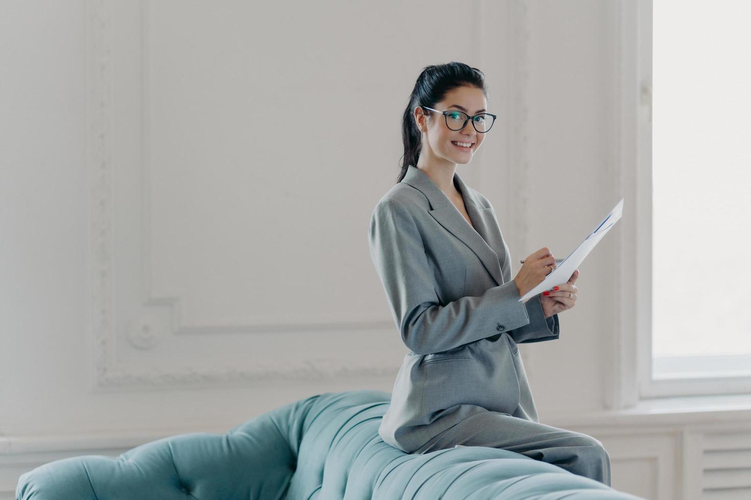 Pleased prosperous woman financial manager studies documents in spacious home office, leans at sofa, dressed in elegant clothes, looks gladfully at camera, writes information. Paper work concept photo