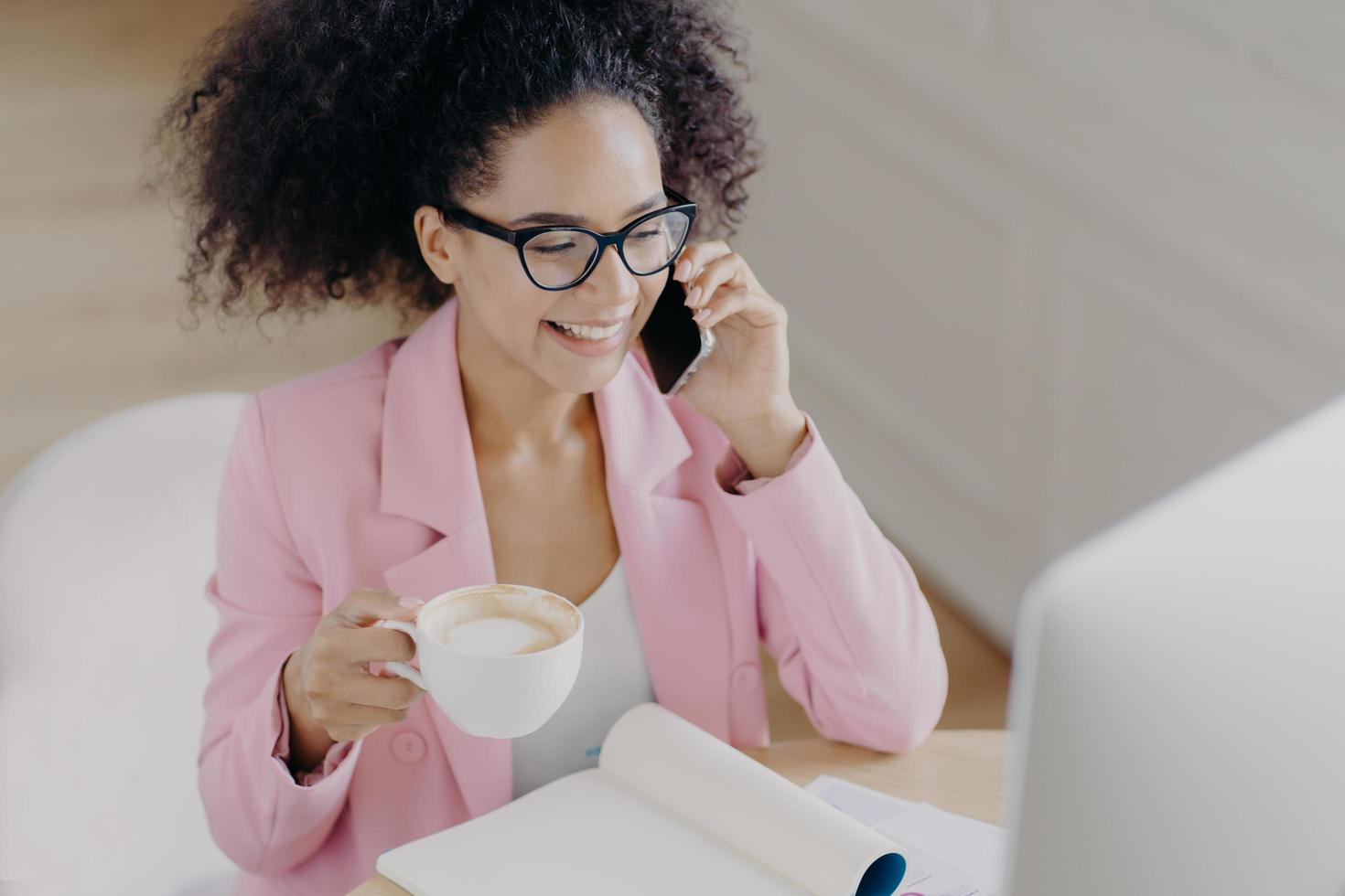 Top view of glad dark skinned woman with curly dark hair, has phone conversation, holds mug of drink, smiles pleasantly while looks at screen of computer, wears elegant attire, busy working at project photo