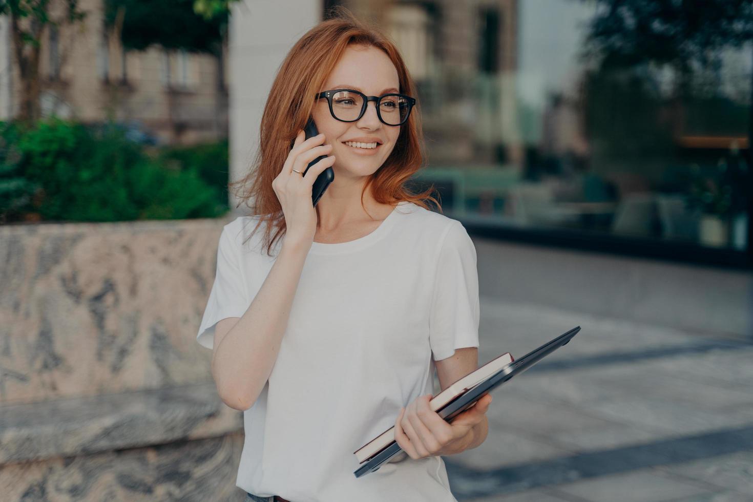 Happy ginger woman has telephone conversation looks into distance holds tablet and notebook photo