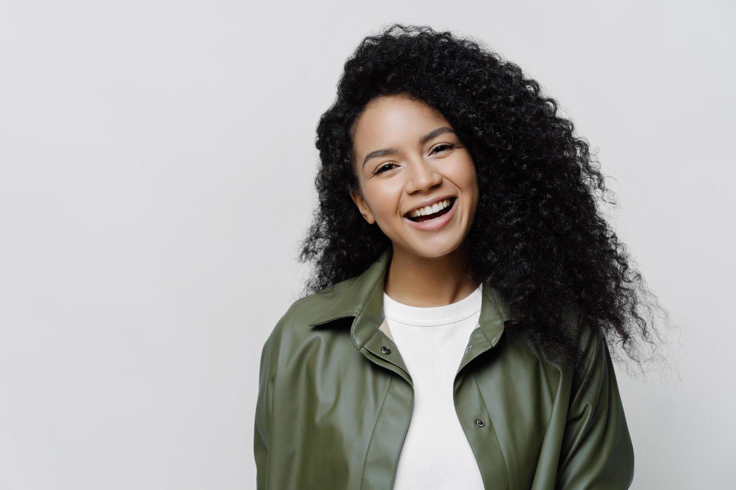 Portrait of carefree joyful African American lady with beaming smile and curly bushy hair, glad to be promoted at work, hears excellent news, dressed in leather shirt, isolated on white background photo