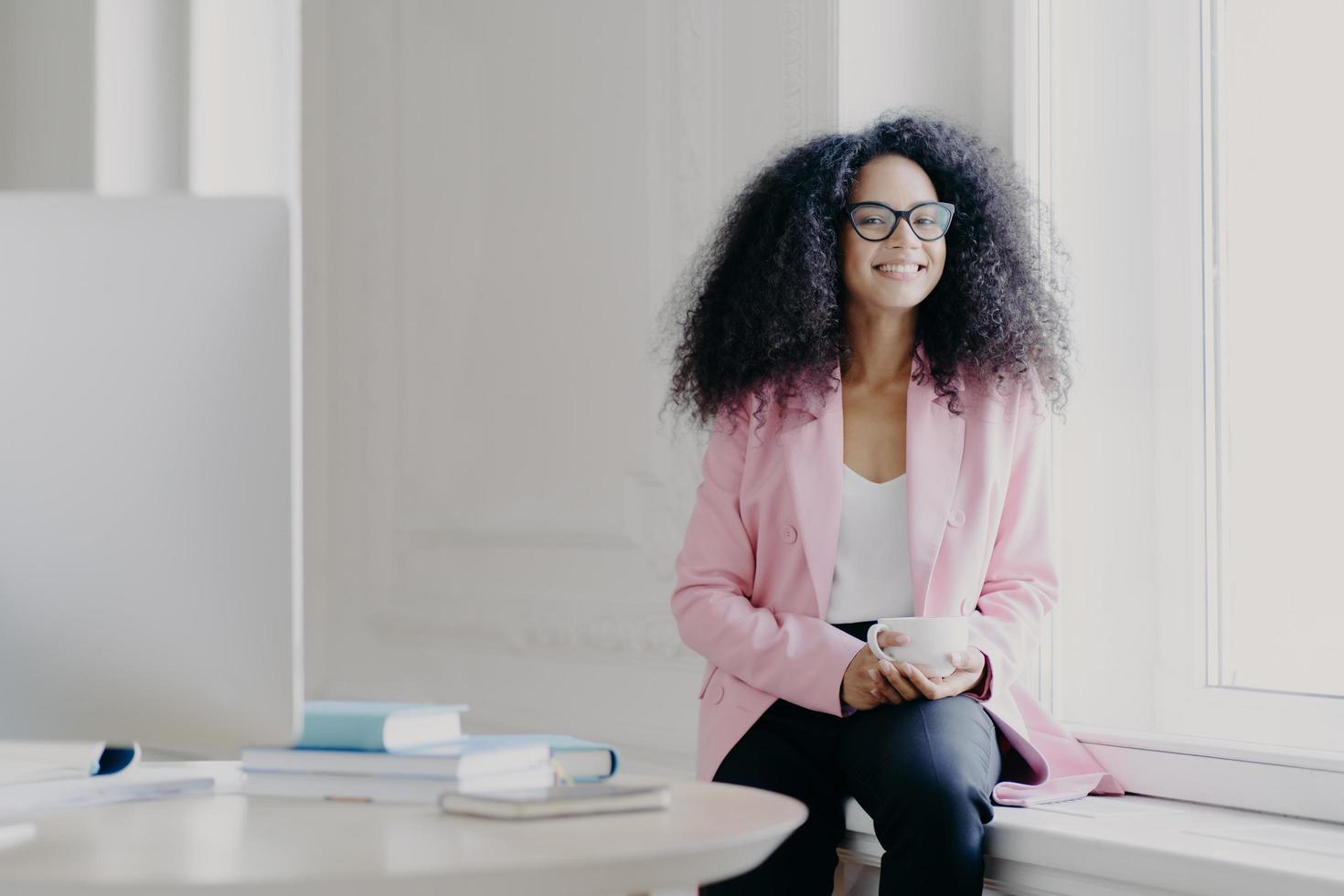 Happy female entrepreneur has bushy Afro hair, wears rosy jacket and black trousers, holds cup of drink, poses at windowsill in spacious cabinet with table, modern computer, enjoys aromatic coffee photo