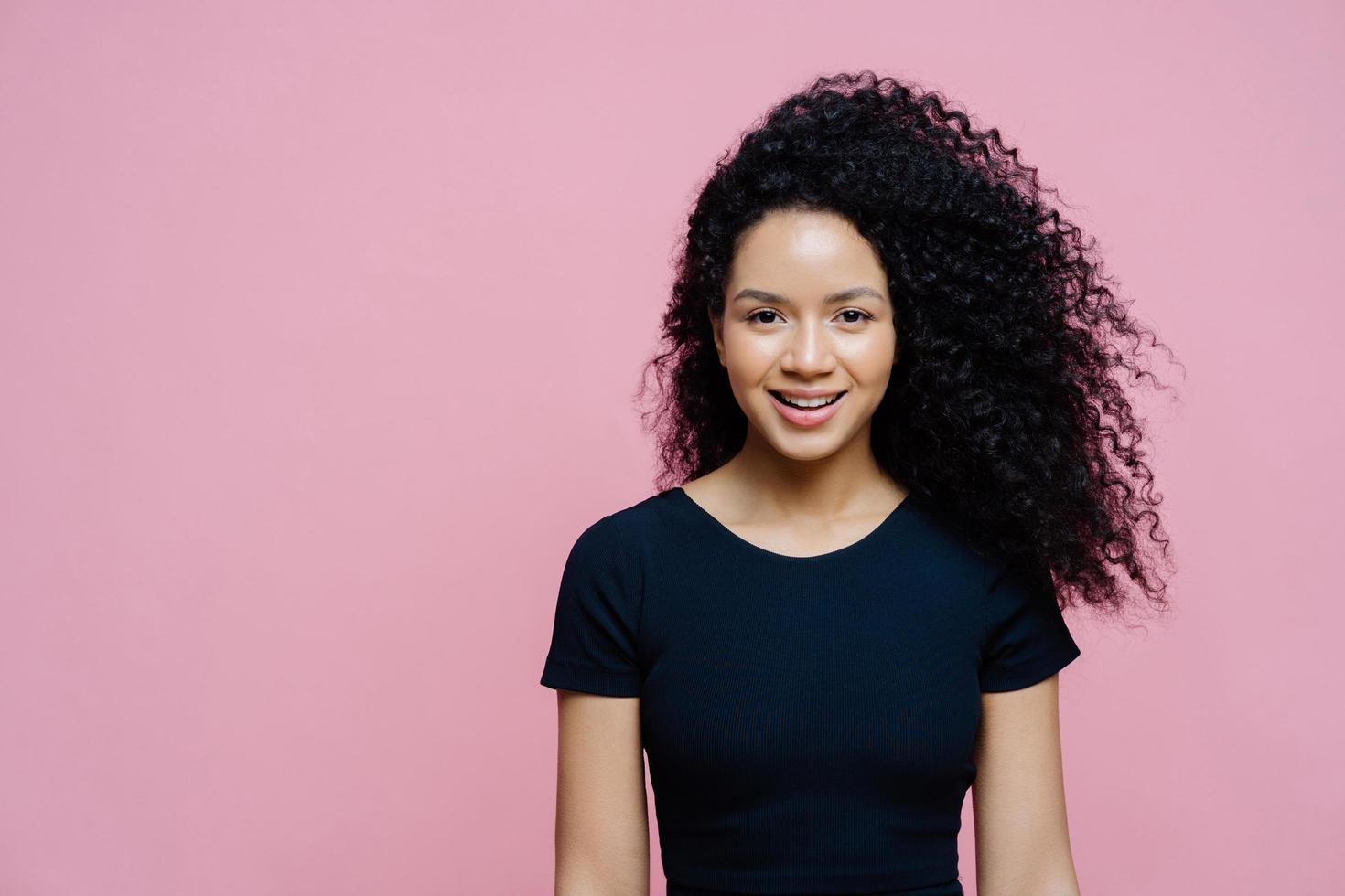 Portrait of beautiful charming dark skinned woman smiles happily, pleased to hear pleasant news, wears casual black t shirt, models against rosy background, copy space aside for your promotion photo