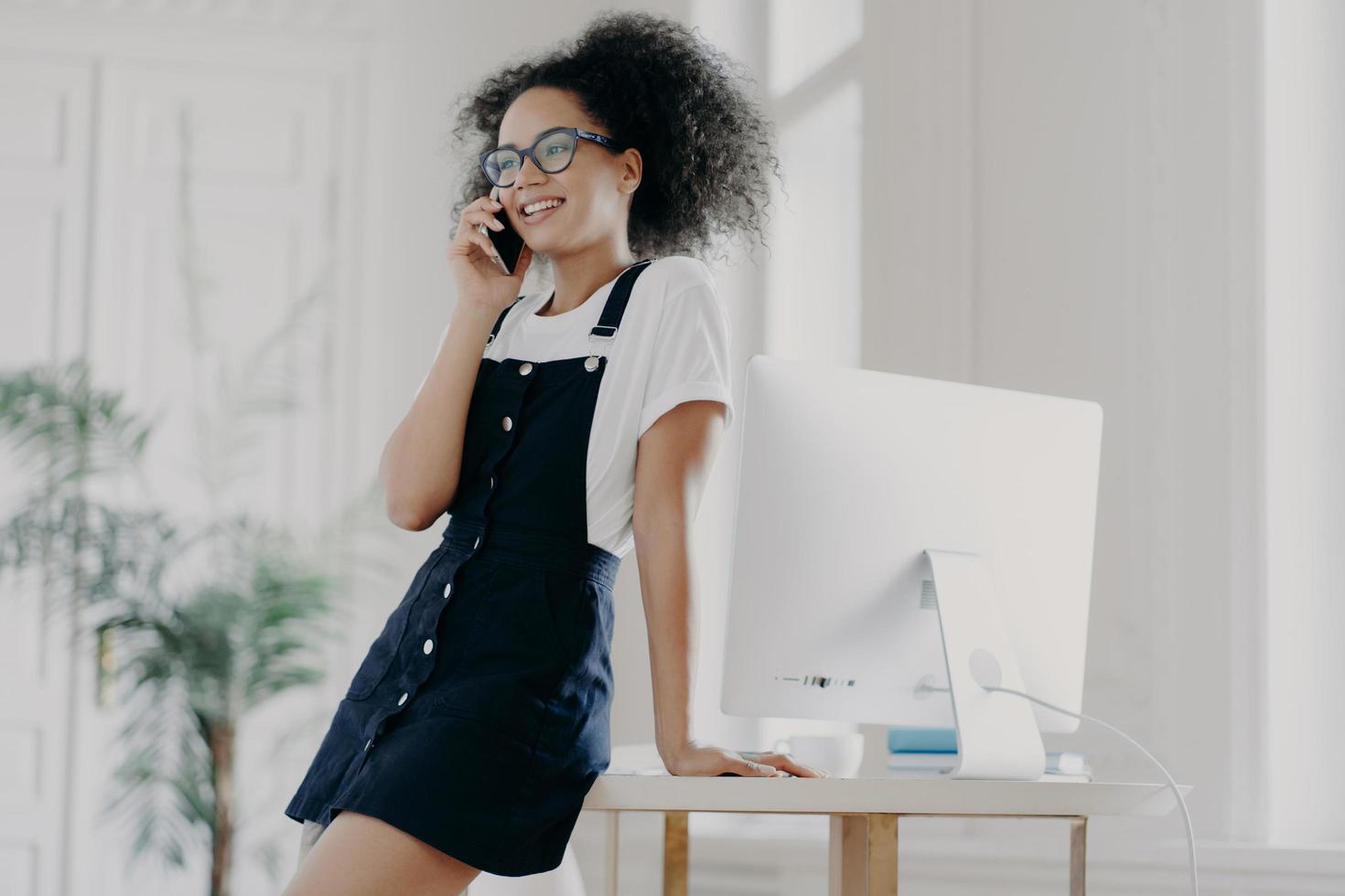 Pleased curly female office worker dressed in black sarafan, leans at table, poses in cabinet near computer monitor, has telephone conversation, discusses details of contract, has smile on face photo